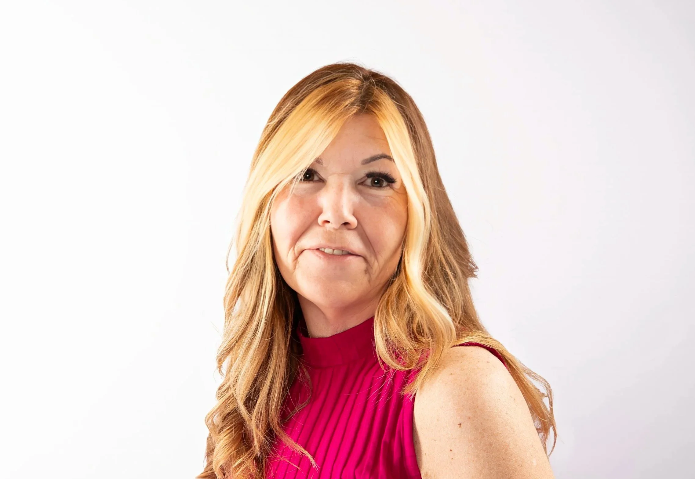 A woman with long, wavy blonde hair and a side part, wearing a sleeveless, bright pink top, posing against a white background.