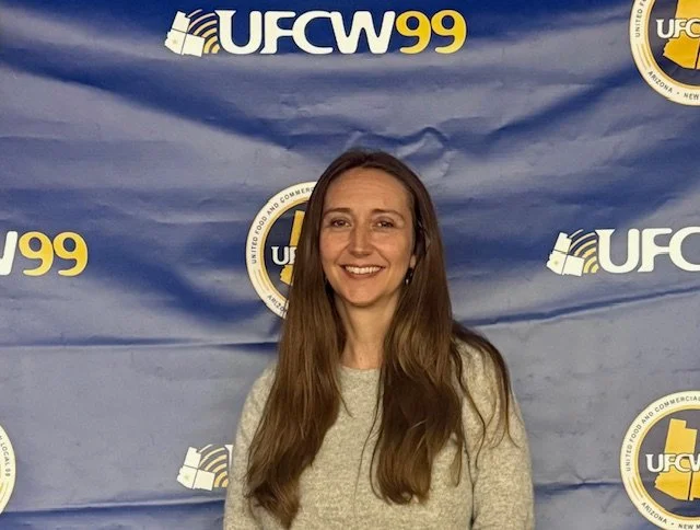 A woman with long brown hair smiling in front of a blue UFCW 99 banner.