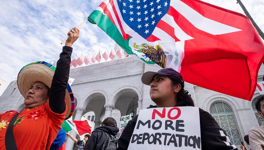 Protesters demonstrating with flags and signs outside a government building, advocating against deportation.