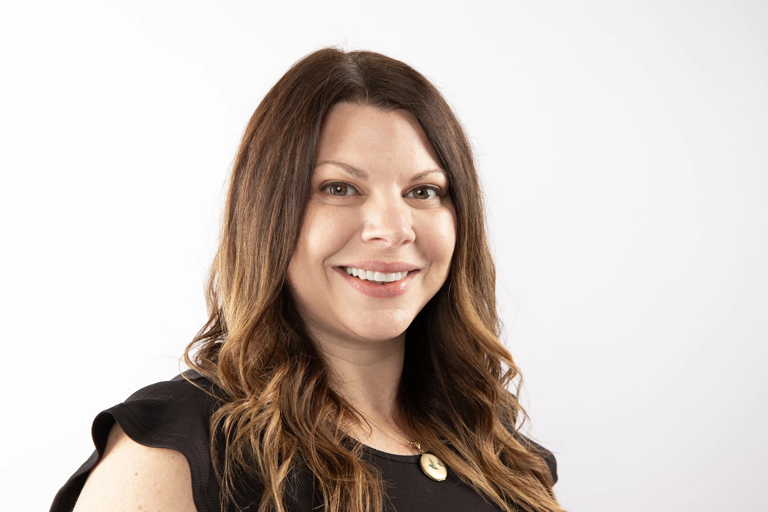 A woman with long wavy brown hair smiling, wearing a black sleeveless top and a round pendant necklace, against a plain white background.