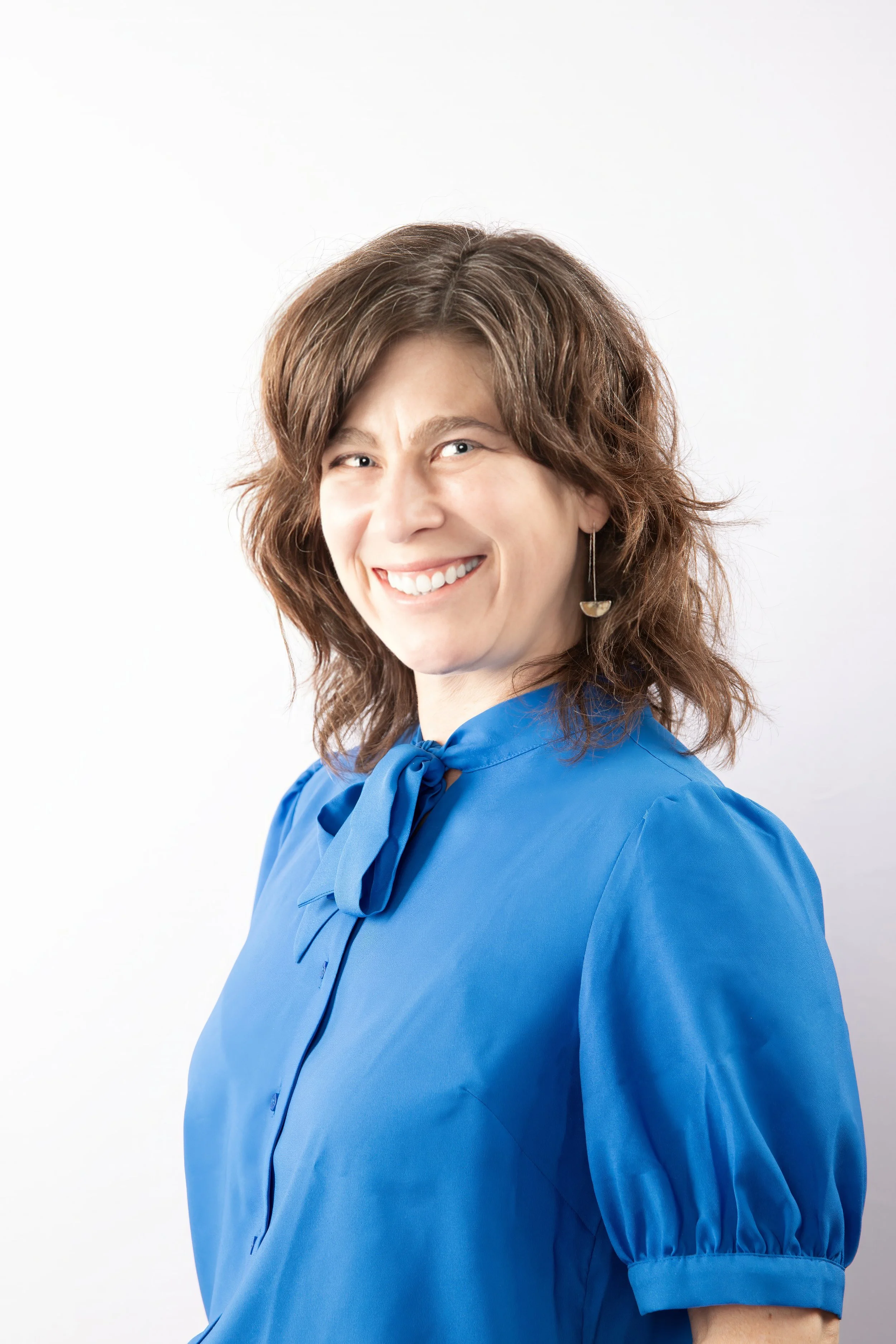 A woman with medium-length wavy brown hair smiling, wearing a bright blue blouse with a bow tie at the collar, and an earring with a half-moon shape, against a plain white background.