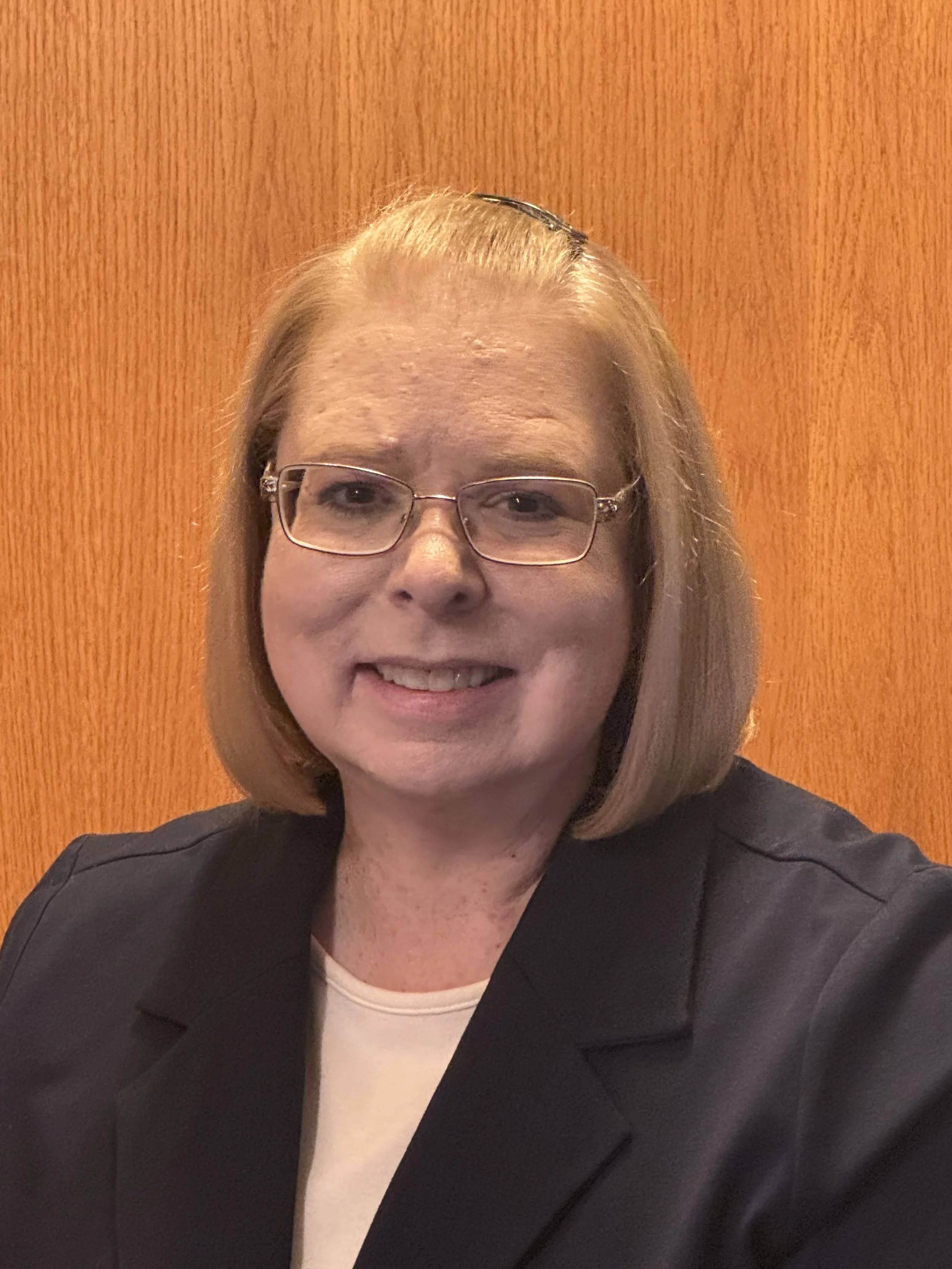 A woman with shoulder-length blonde hair, glasses, wearing a black blazer and a light-colored top, smiling in front of a wooden background.