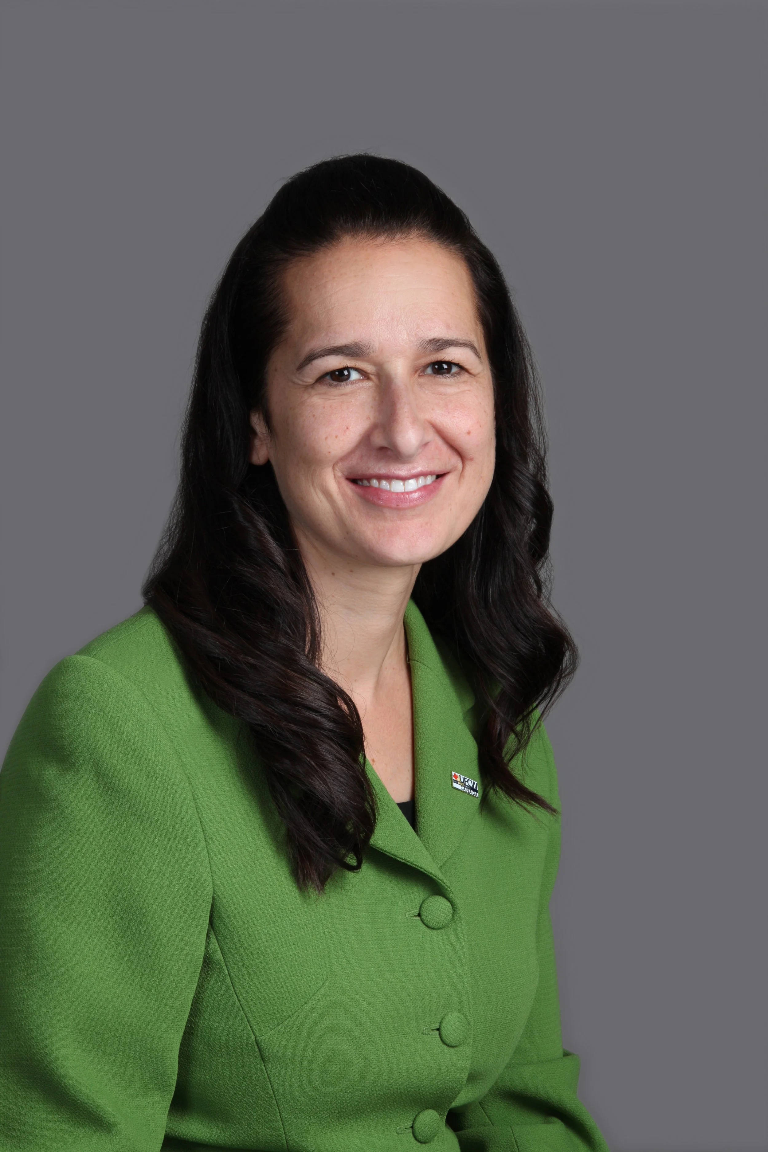 Professional woman with long dark hair, wearing a green blazer, smiling against a gray background.