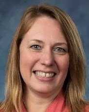 Close-up of a woman smiling with medium-length blonde hair, wearing a maroon top, against a blue background.