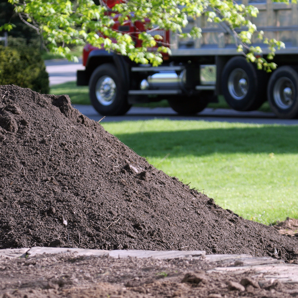 Large pile of Screened Topsoil, with a green lawn and a tree with green leaves, and a red truck with a flatbed trailer in the background.