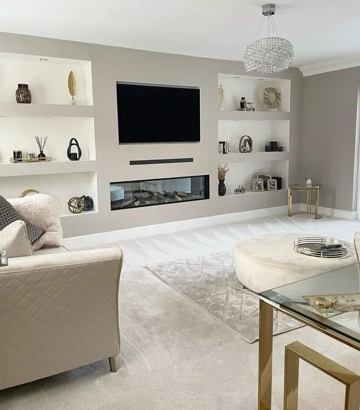 Modern living room with built-in shelves, a wall-mounted TV, a gas fireplace below the TV, a chandelier, a white armchair, an oval ottoman, a glass coffee table with gold accents, and decorative items on shelves.