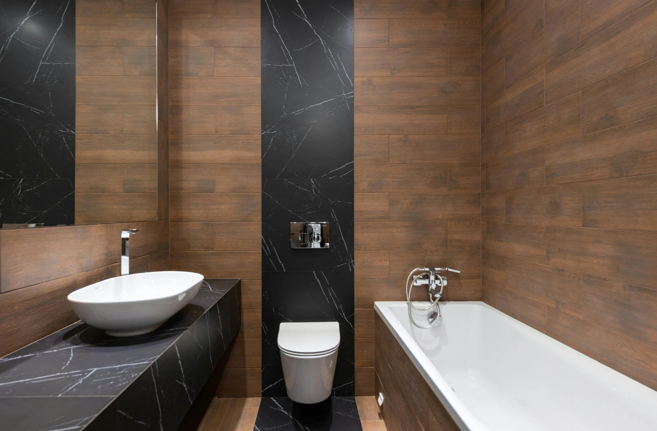 Modern bathroom with wood-textured walls, black marble accents, a white vessel sink with a chrome faucet, and a white bathtub with a wall-mounted faucet and handheld shower head.