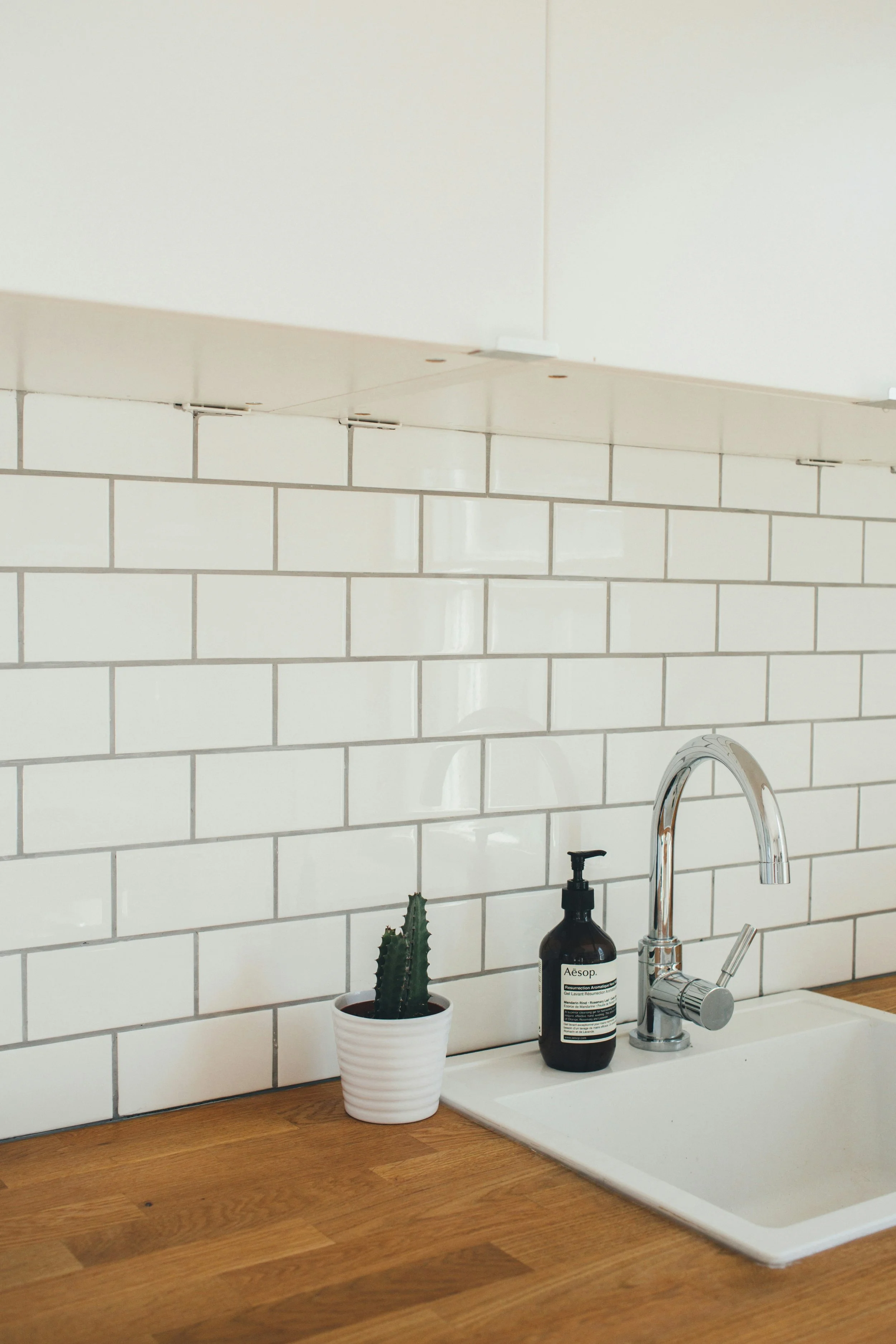A kitchen countertop with a white ceramic sink, a modern chrome faucet, a potted cactus plant, and a bottle of liquid soap against a background of white subway tiles.
