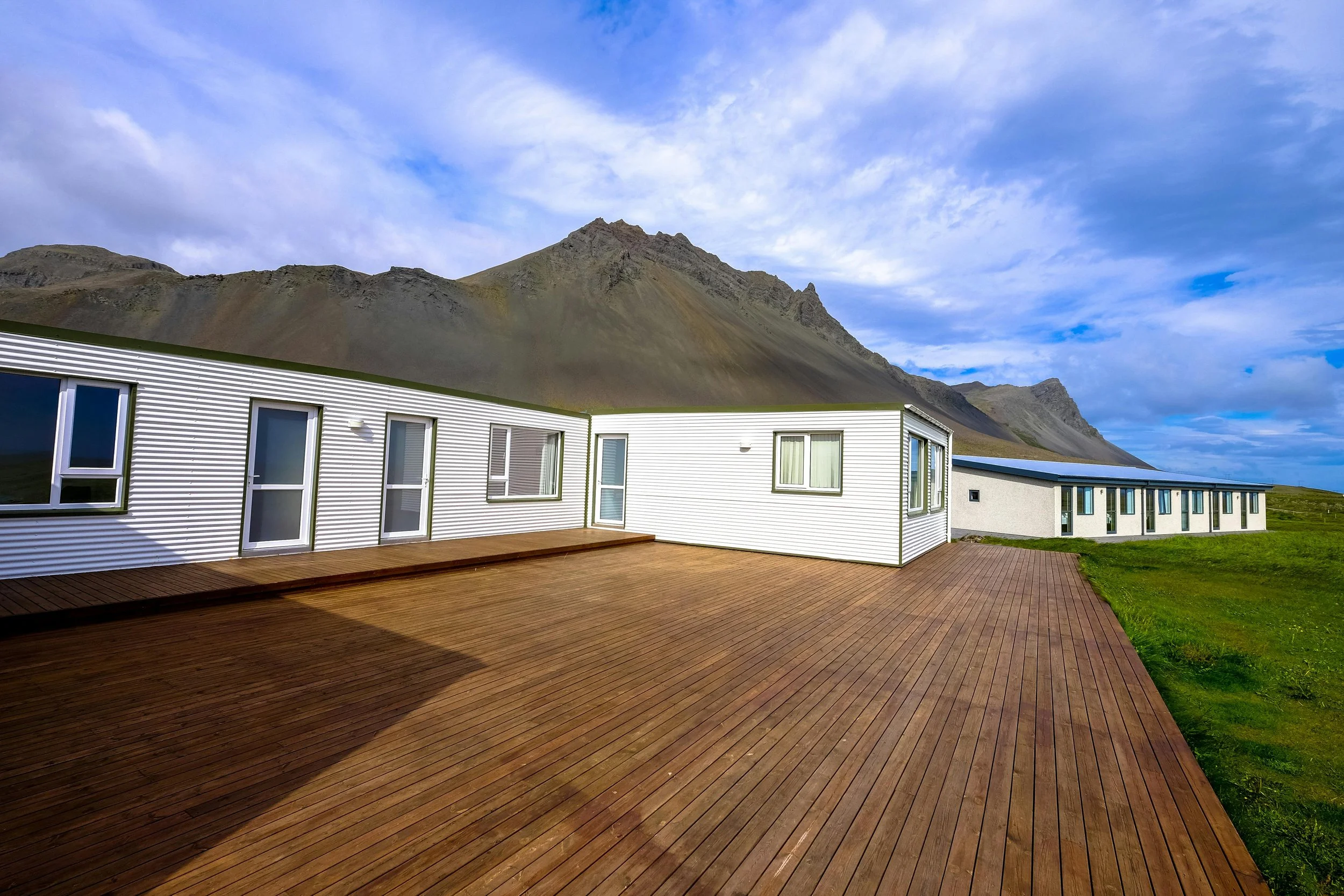Modern white building with multiple windows and a wooden deck in front, set against a mountainous landscape with a cloudy sky.