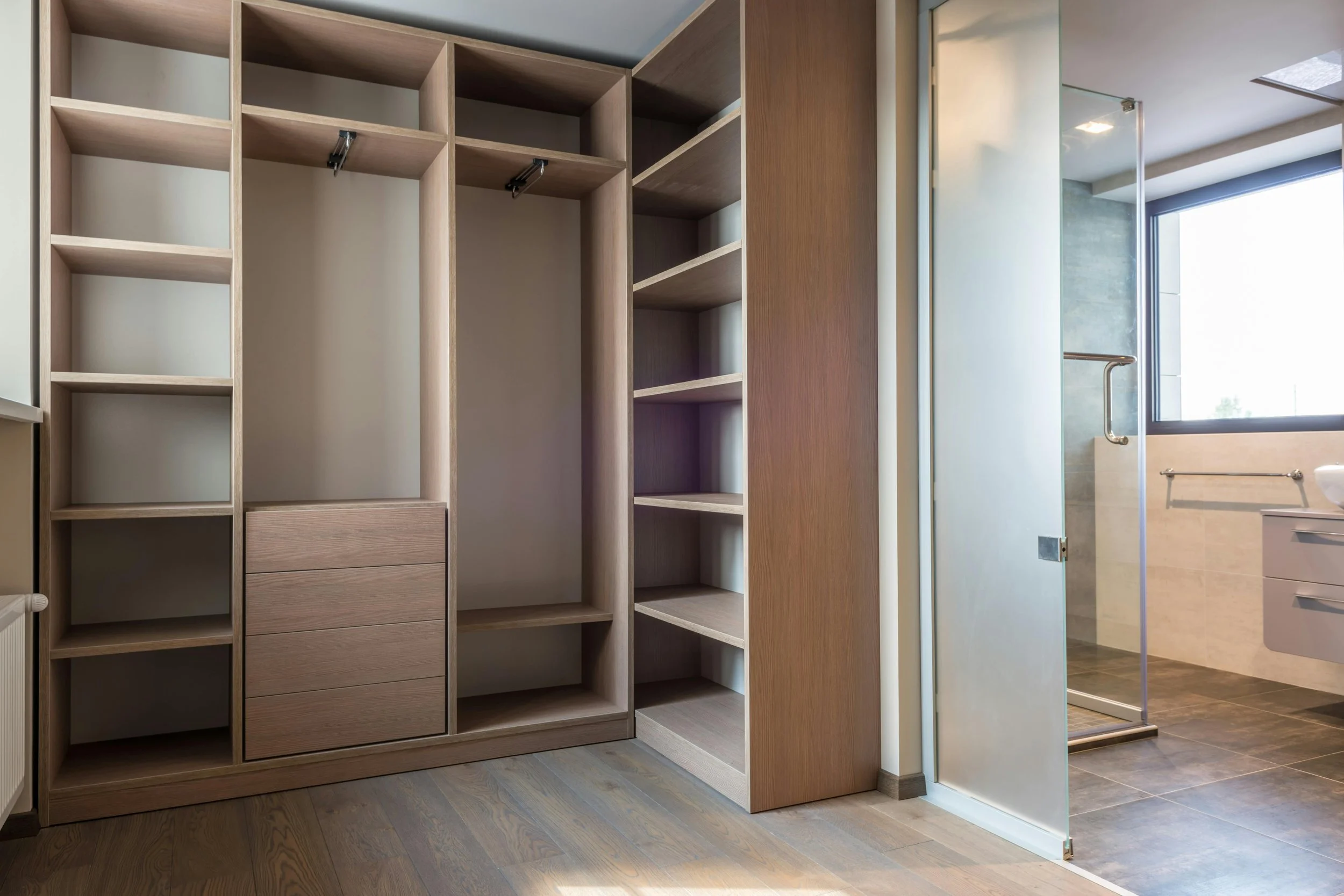 Empty wooden closet with shelves and drawers next to a bathroom with a glass shower door, large window, and gray tile floor.