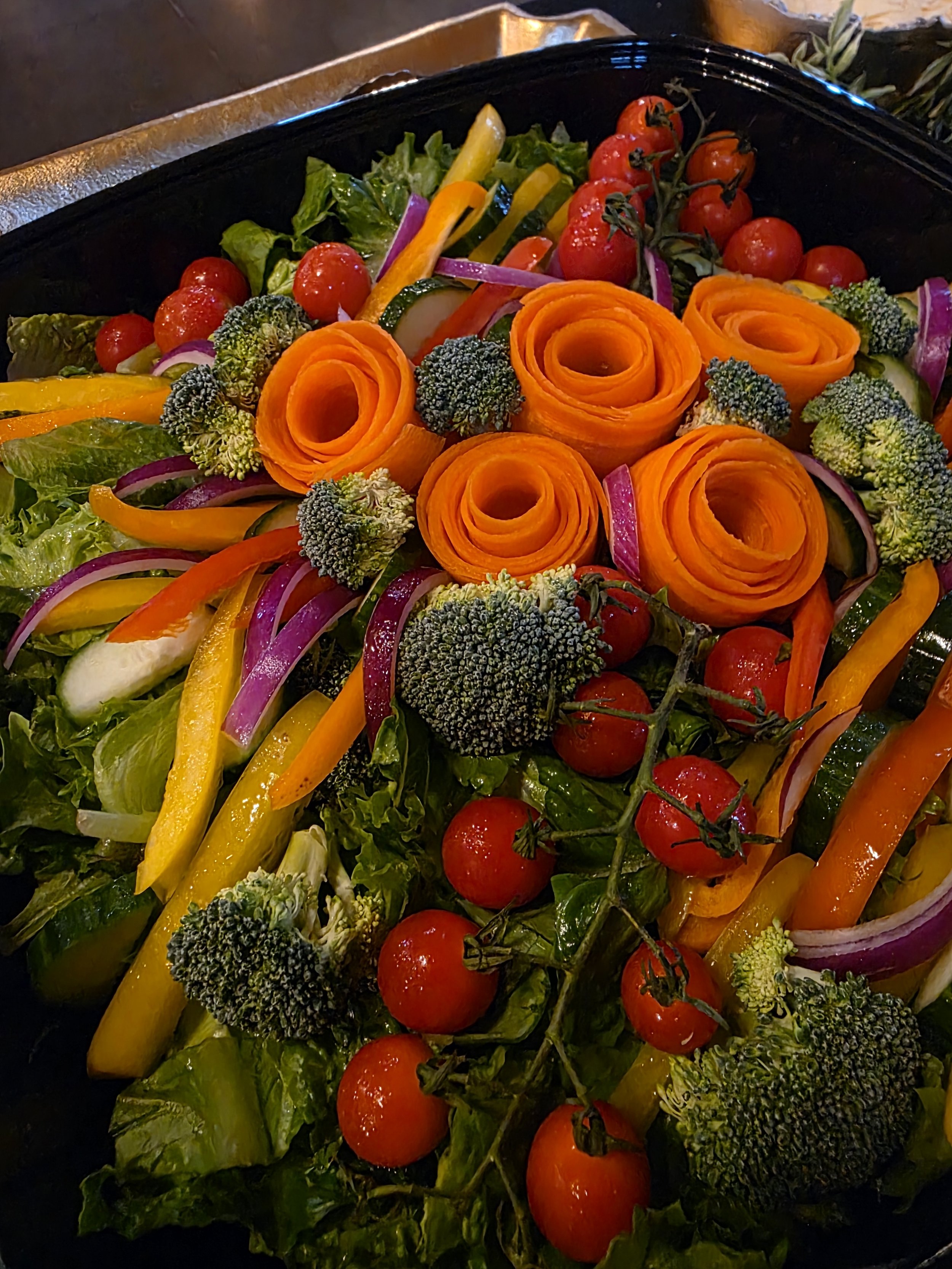 Fresh vegetable platter with cherry tomatoes, broccoli, sliced cucumbers, red onion, bell peppers, and carrot roses on a black tray.