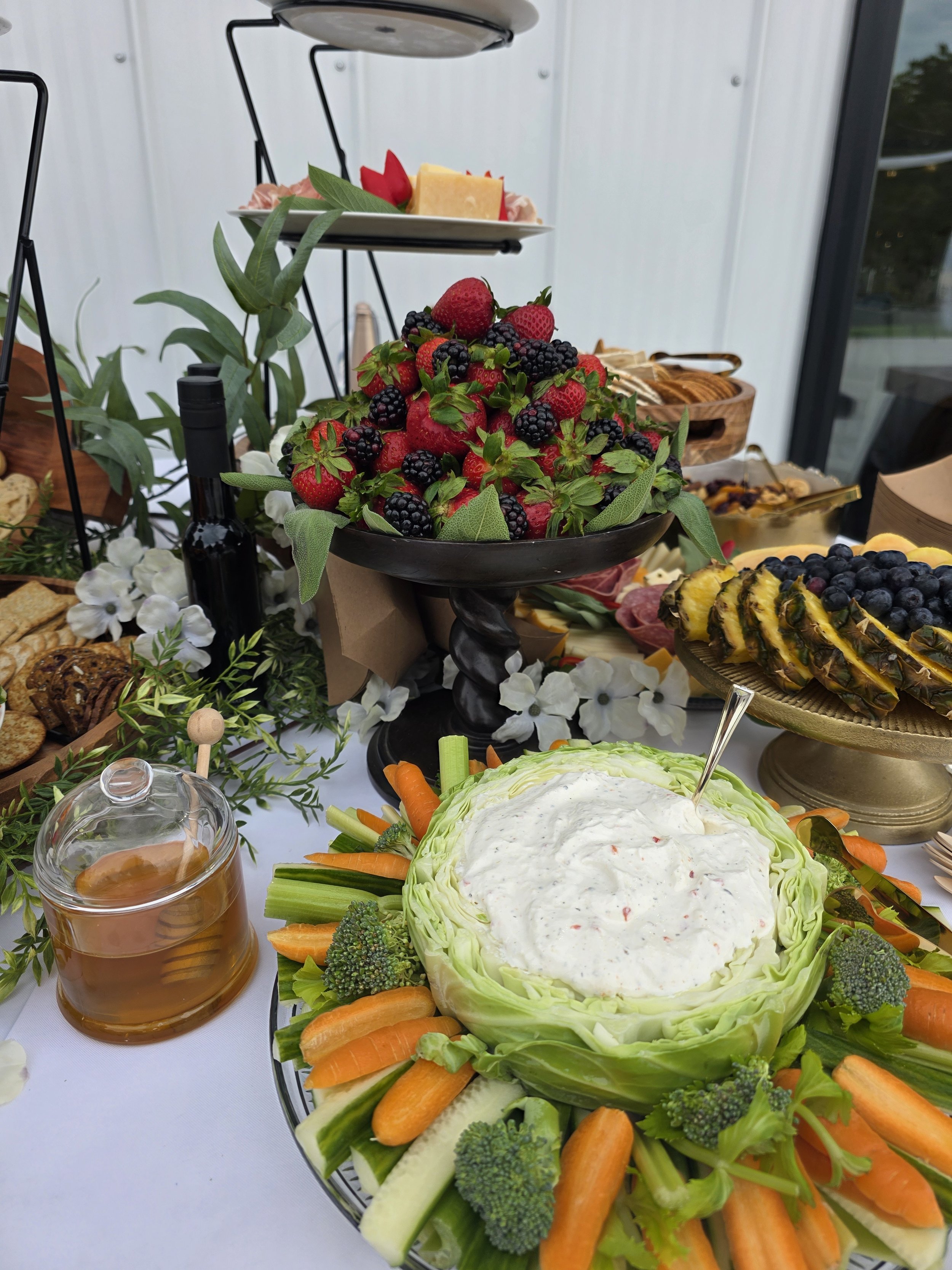 A buffet table with fresh fruit, vegetables, cheeses, and other appetizers, including strawberries, blackberries, pineapple, carrots, broccoli, and lettuce, arranged for a gathering.