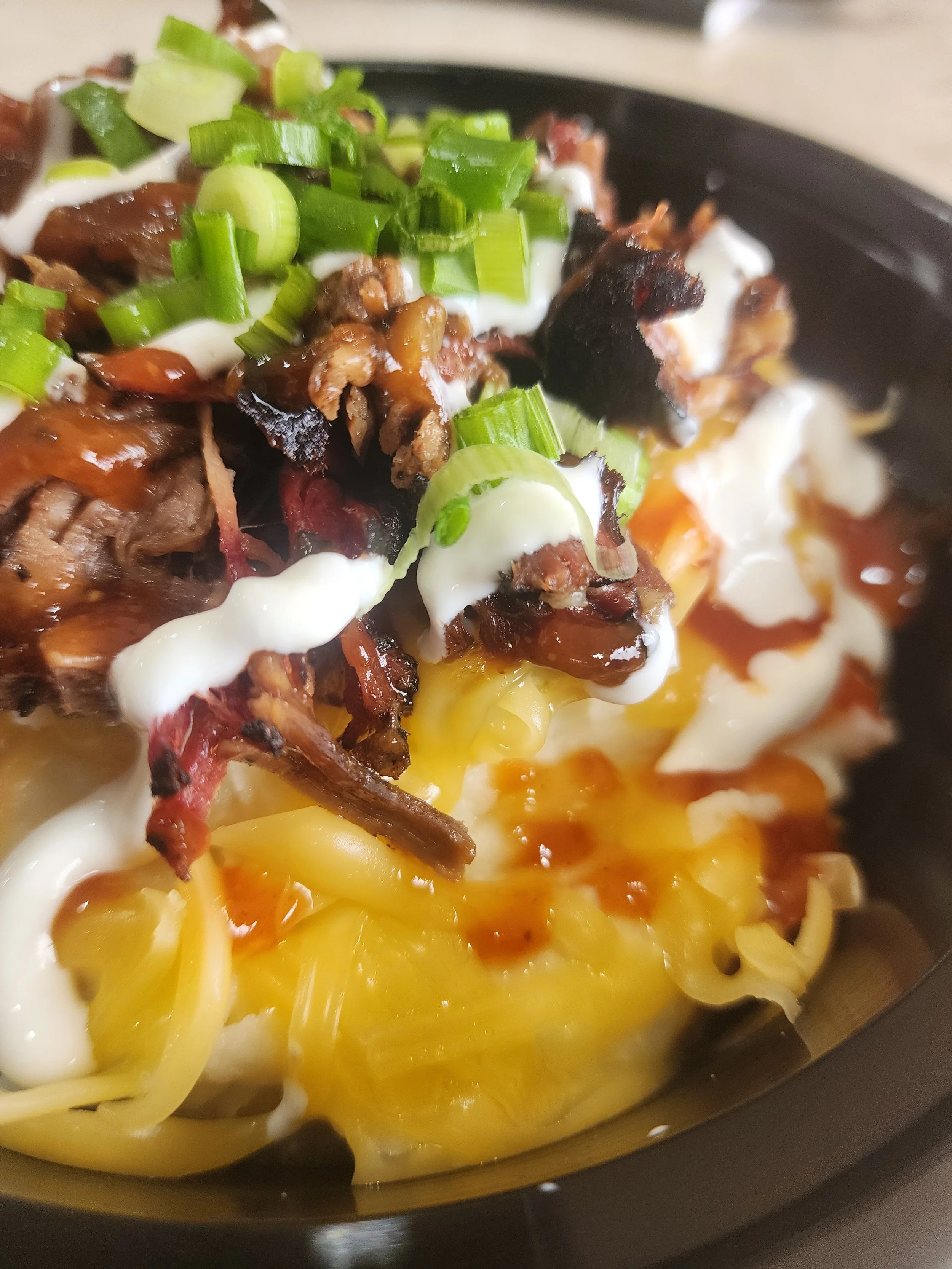 Close-up of a dish with melted cheese, chopped green onions, and cooked meat with barbecue sauce on a black plate.