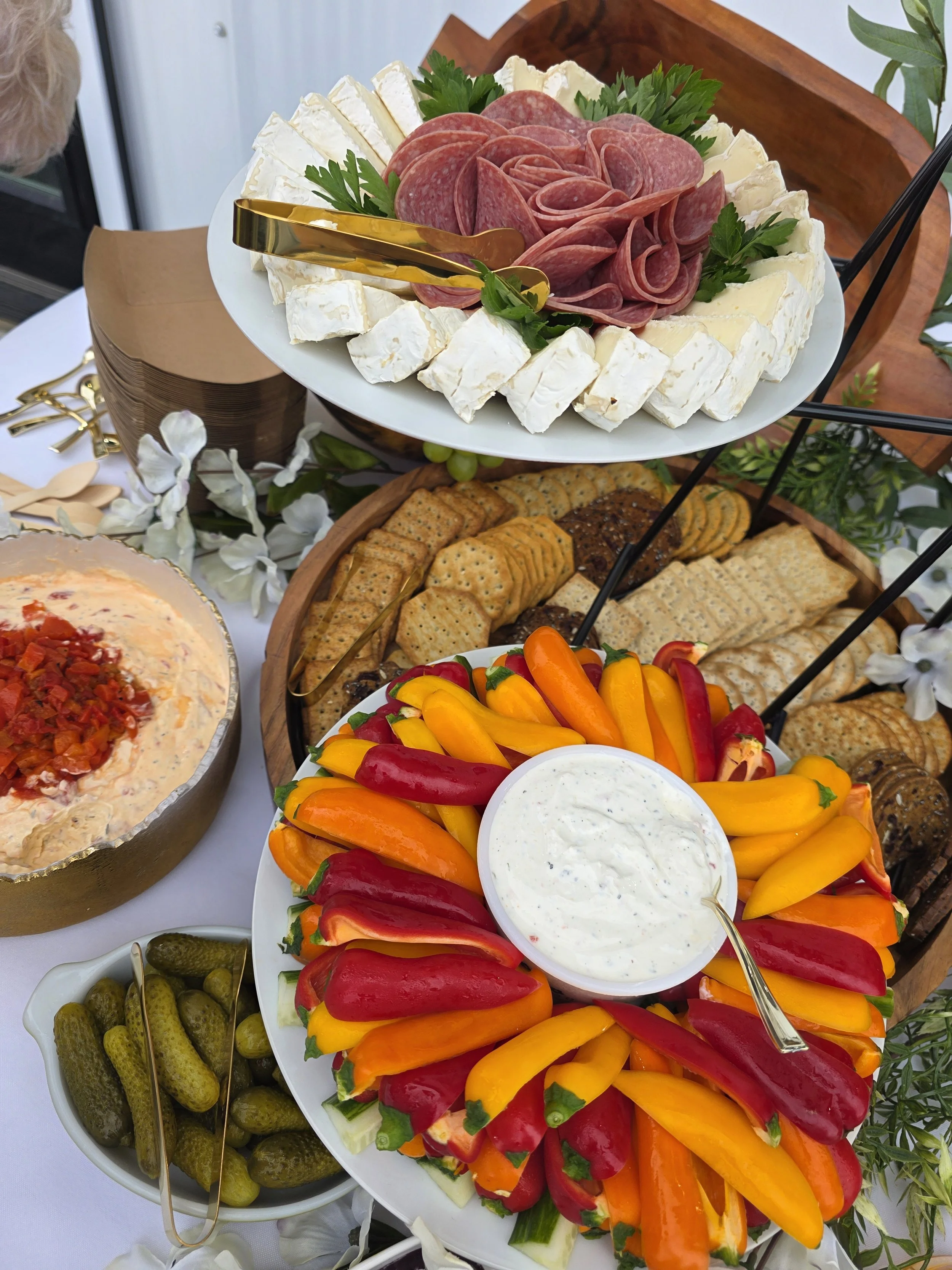 A food platter featuring sliced cheese, various cured meats, fresh vegetables including cherry tomatoes, colorful bell peppers, and a bowl of creamy dip or dressing, arranged on a tiered serving tray with side bowls of pickles and olives.