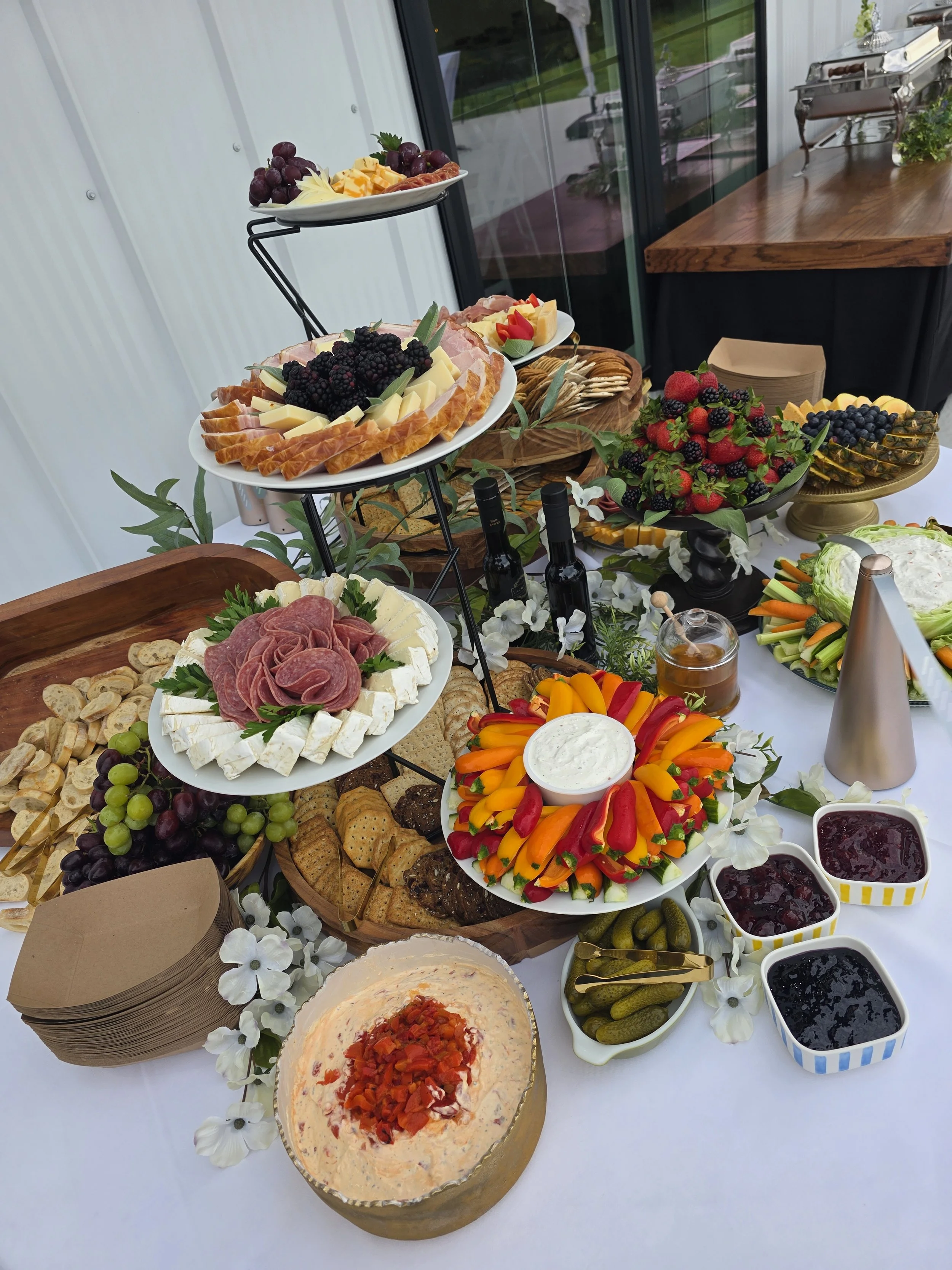 A table with various appetizers, cheeses, fruits, vegetables, spreads, and condiments at a buffet or party setting, featuring a cheese and meat platter, fresh fruit bowl, vegetable platter, and small bowls of jams and spreads.