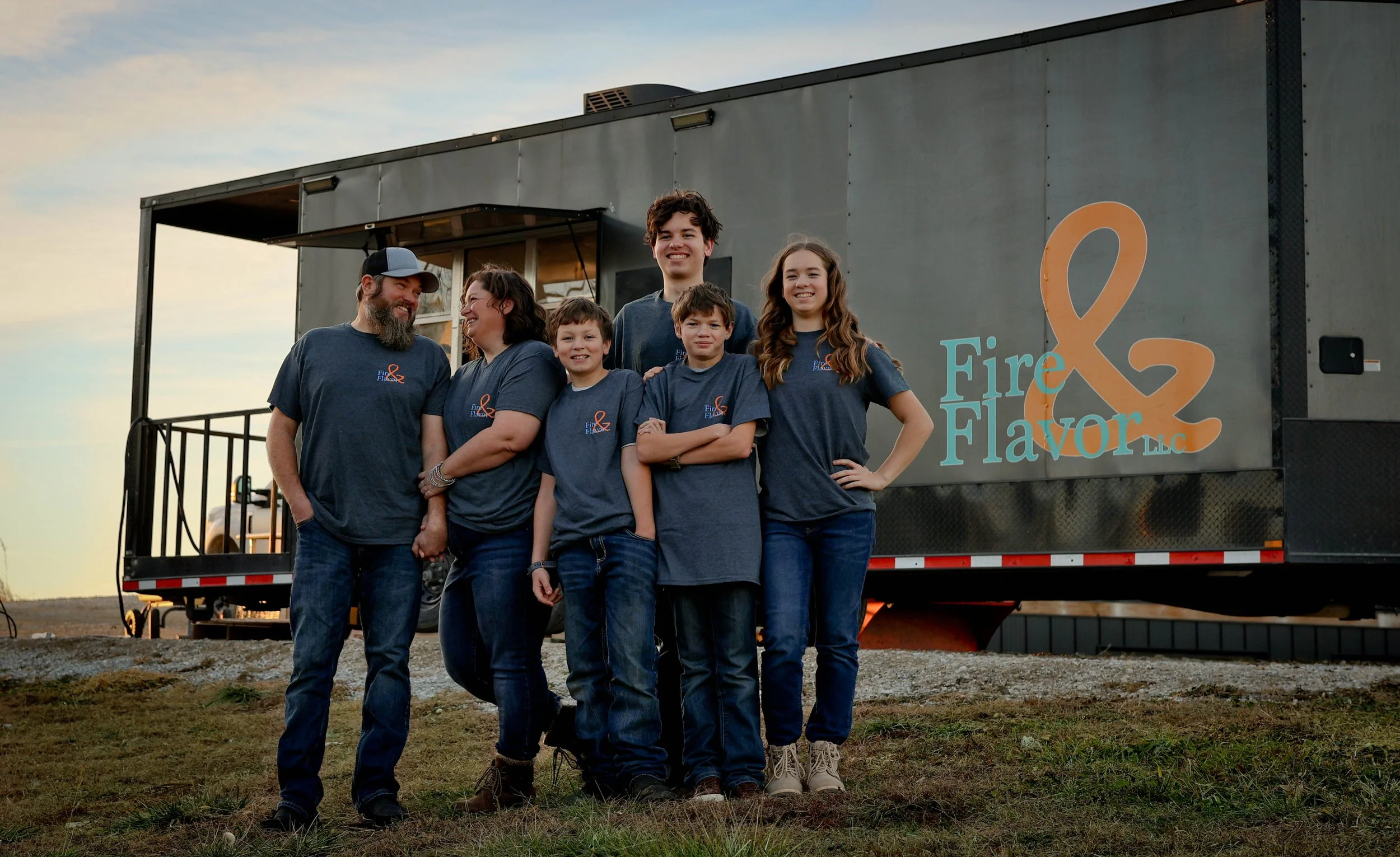 Group of six people standing outdoors in front of a trailer that displays the words "Fire & Flavor LLC" along with an orange ampersand symbol. The group includes three children and three adults, all casually dressed, smiling and posing for the photo during sunset.