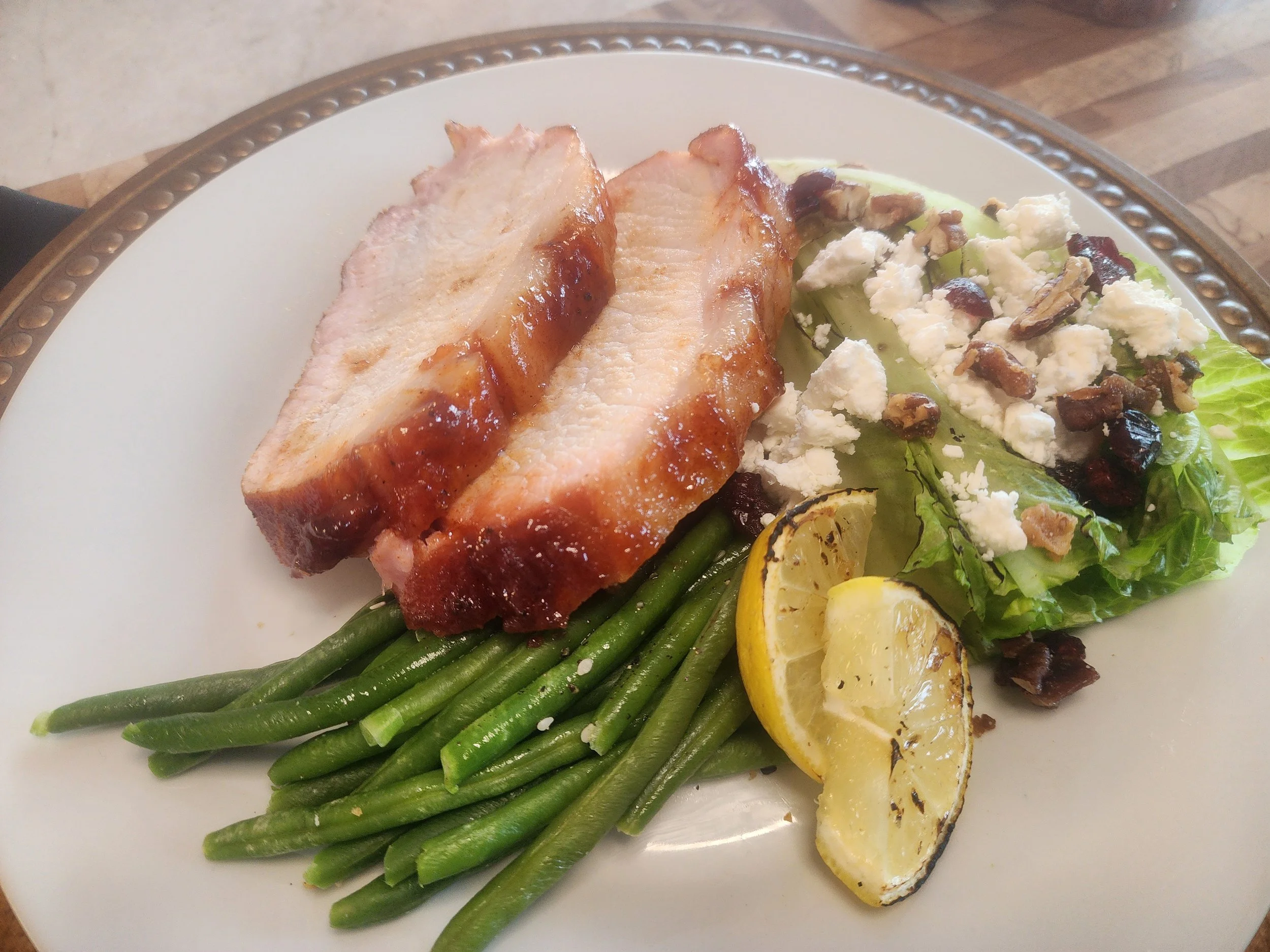 A white plate with two grilled lemon slices, green beans, a grilled chicken breast with sauce, and a salad with lettuce, feta cheese, walnuts, dried cranberries, and sliced almonds.