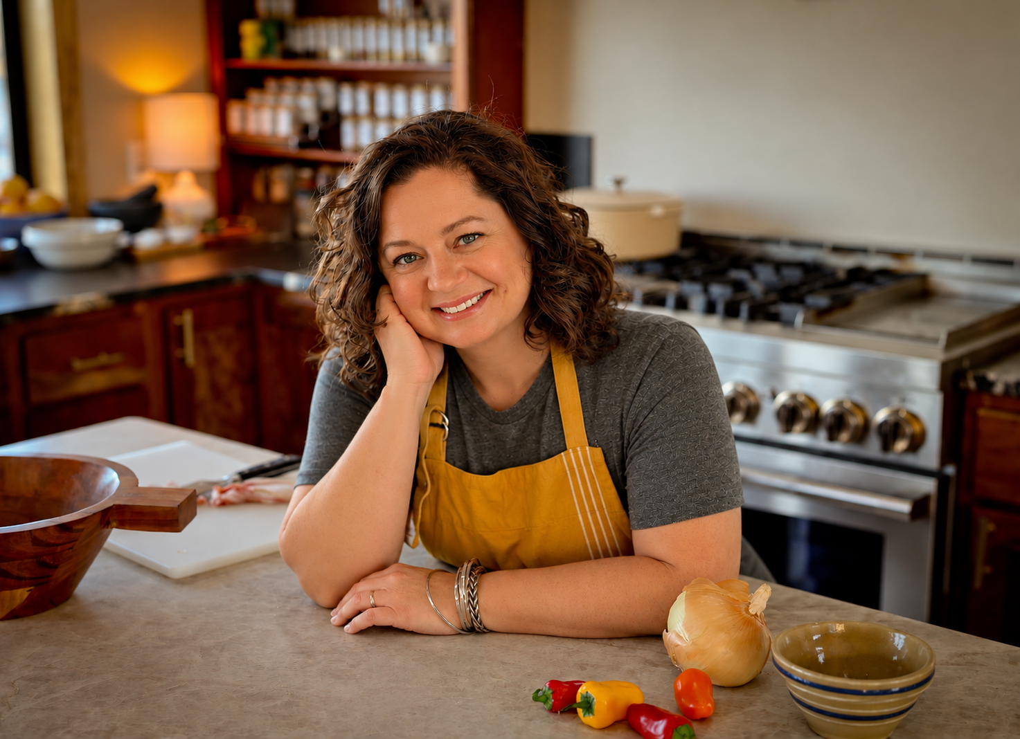 A woman with curly brown hair smiling, resting her head on her hand, standing in a kitchen with a yellow apron, colorful peppers, an onion, a bowl, and a cookbook on the counter.