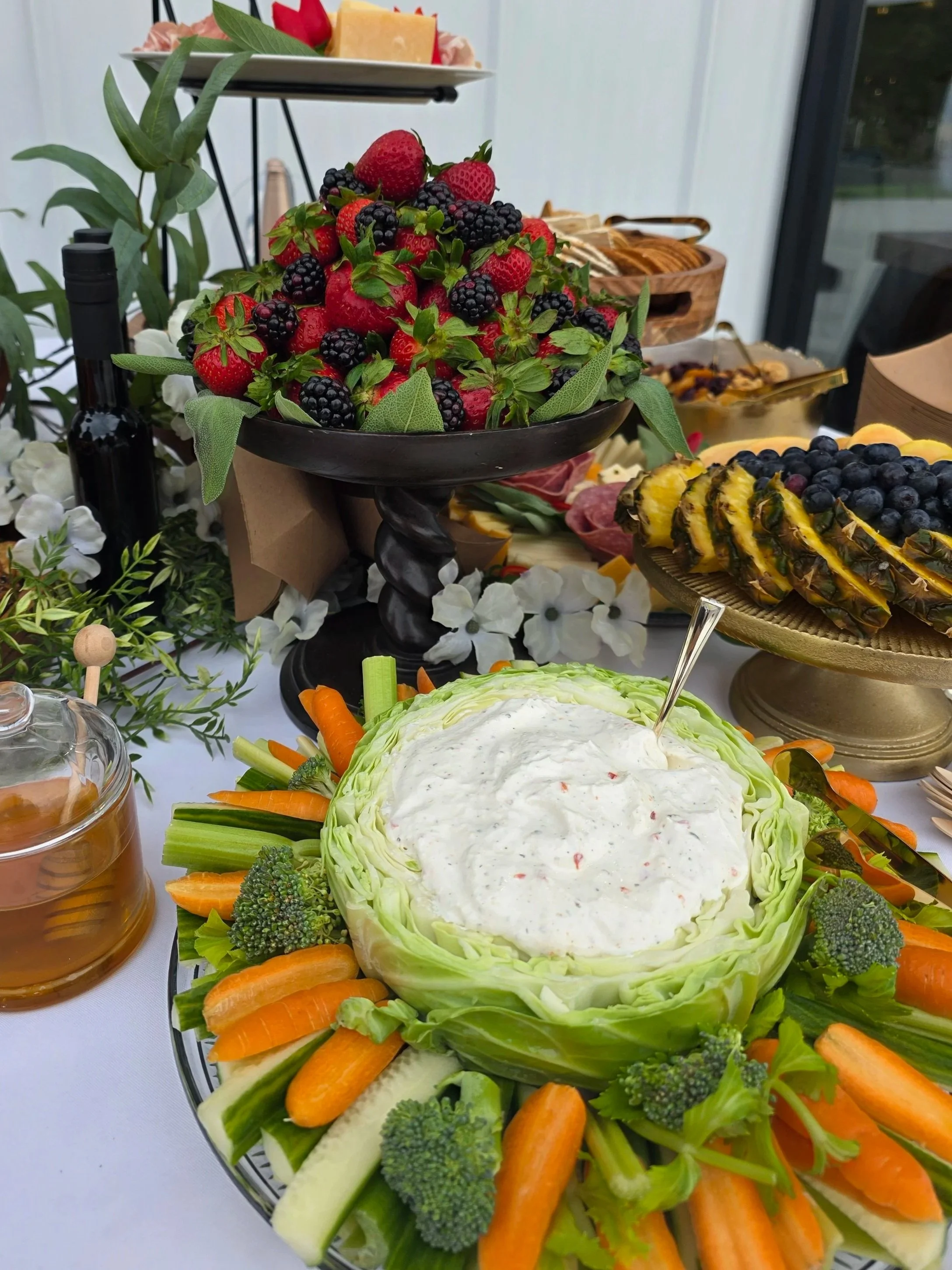 Fresh strawberries and blackberries in a black pedestal bowl, surrounded by sliced carrots, broccoli, cucumber, and lettuce on a silver tray, with a buffet spread of cheese, bread, fruit, and condiments in the background.