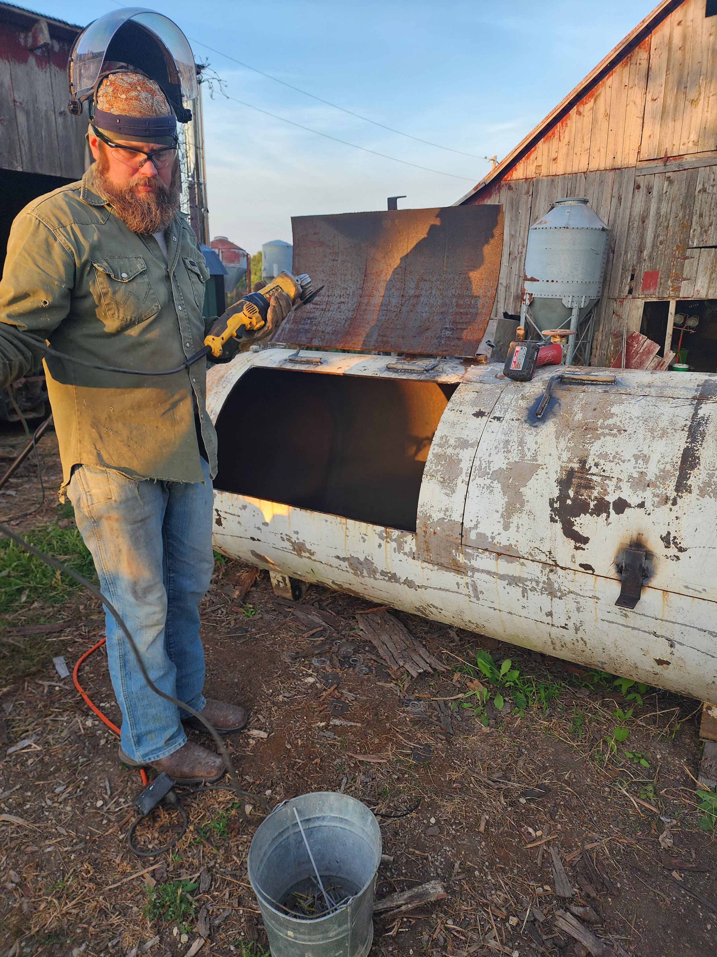 Man welding a custom smoker out of a 500 gallon tank outdoors  and weathered barn with farm equipment in background.