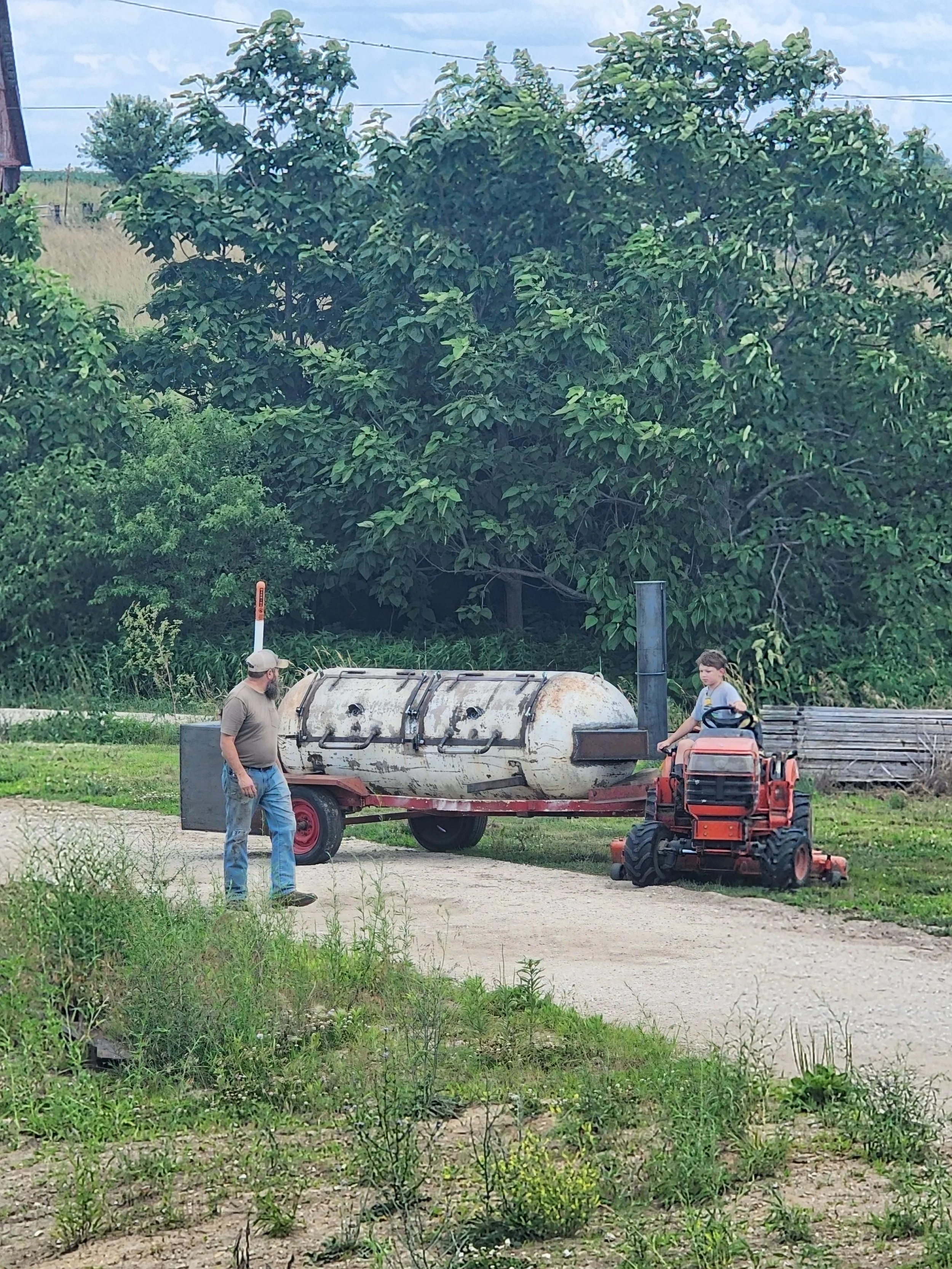 A man and a boy are working outside with gardening or farm equipment. The man stands near a trailer with a custom smoker, while the boy operates a small orange riding mower.