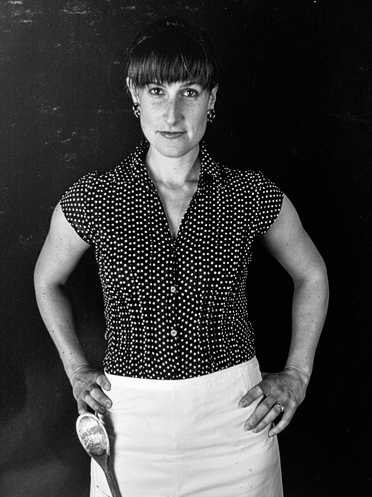 Black and white photo of a woman wearing a polka dot blouse and white skirt, standing against a dark background, holding a large spoon in her right hand.