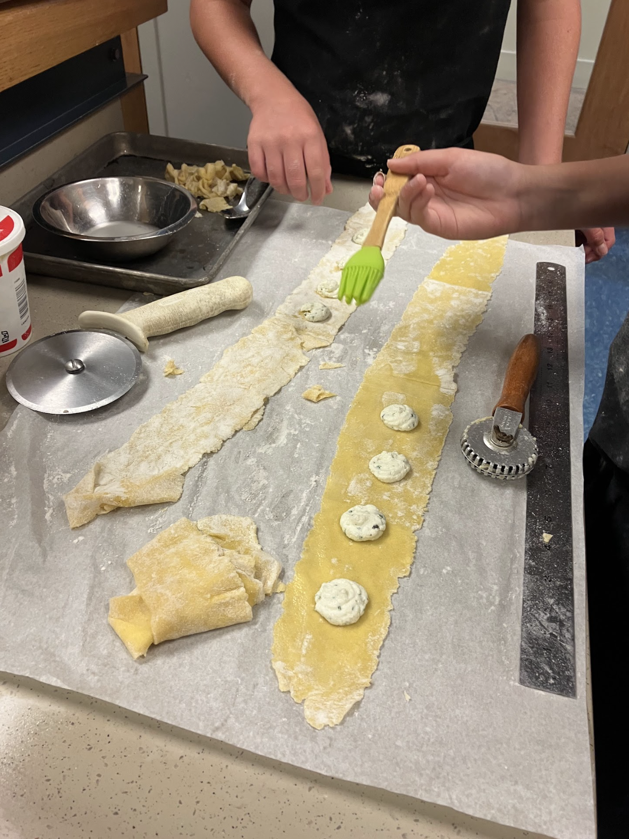 People making fresh pasta with filling, rolling out dough and placing dollops of filling onto the pasta sheet in a kitchen workspace.