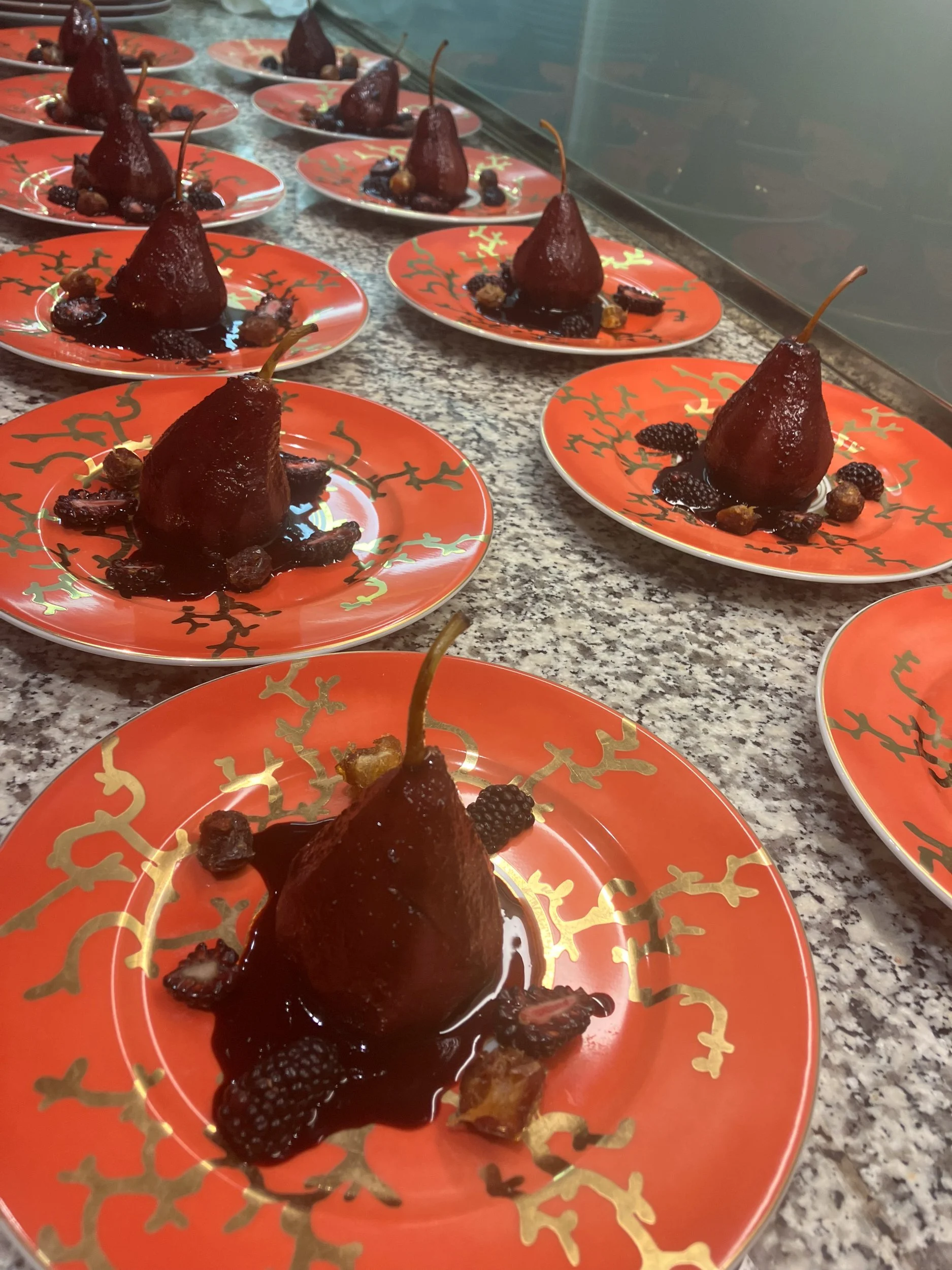 Multiple plates with poached pears topped with sauce and garnished with blackberries and golden raisins on festive red and gold plates.