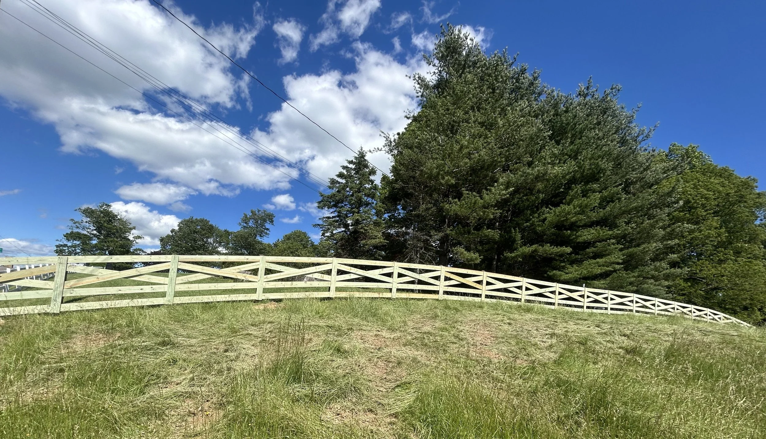 A grassy hill with a white wooden fence running across the middle. In the background, tall green trees and a bright blue sky with scattered white clouds. Power lines are visible crossing the sky.