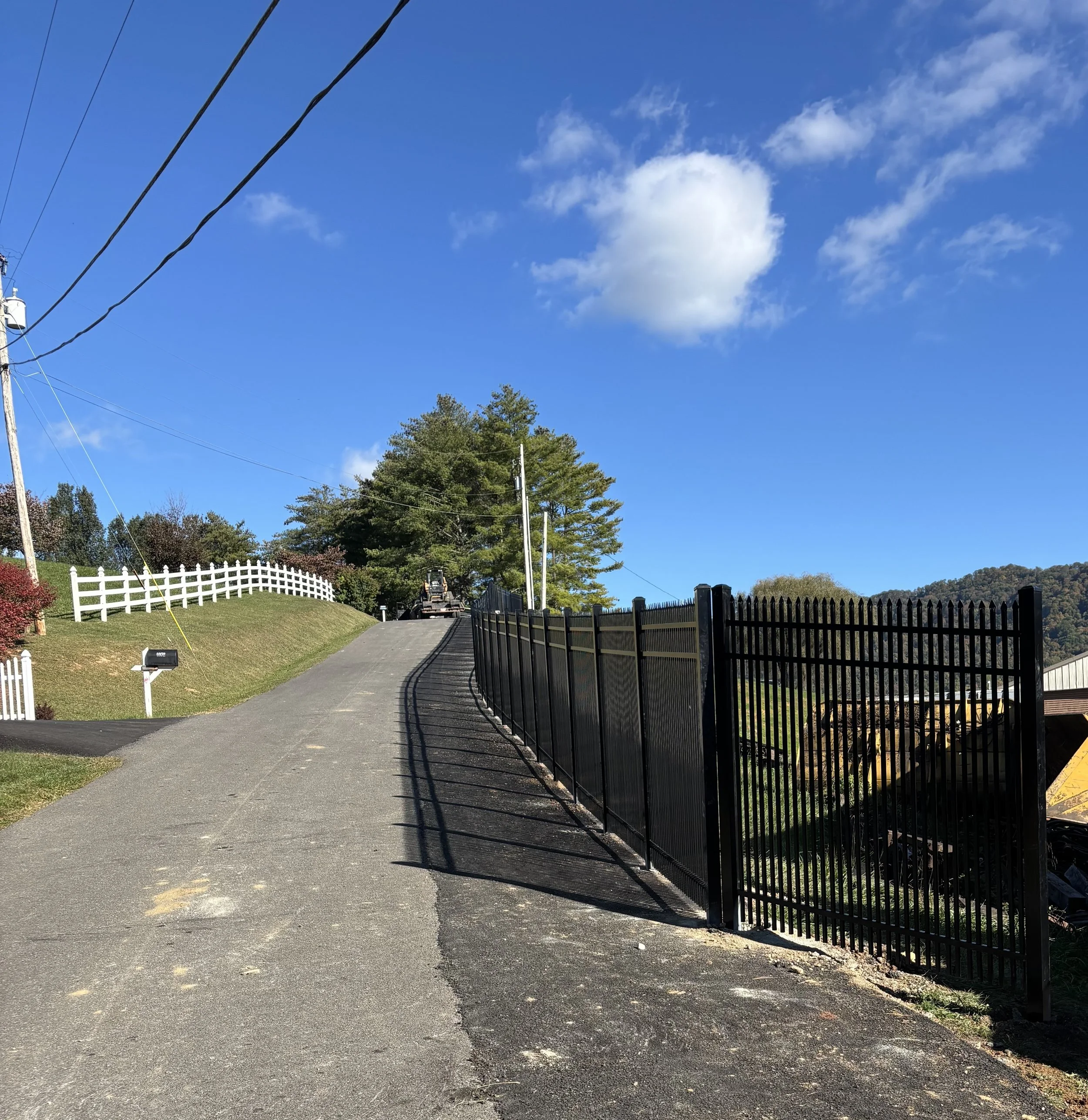 A paved driveway curving uphill with a black metal fence on the right and a white wooden fence on the left. Trees, power lines, and a blue sky with a cloud are visible in the background.