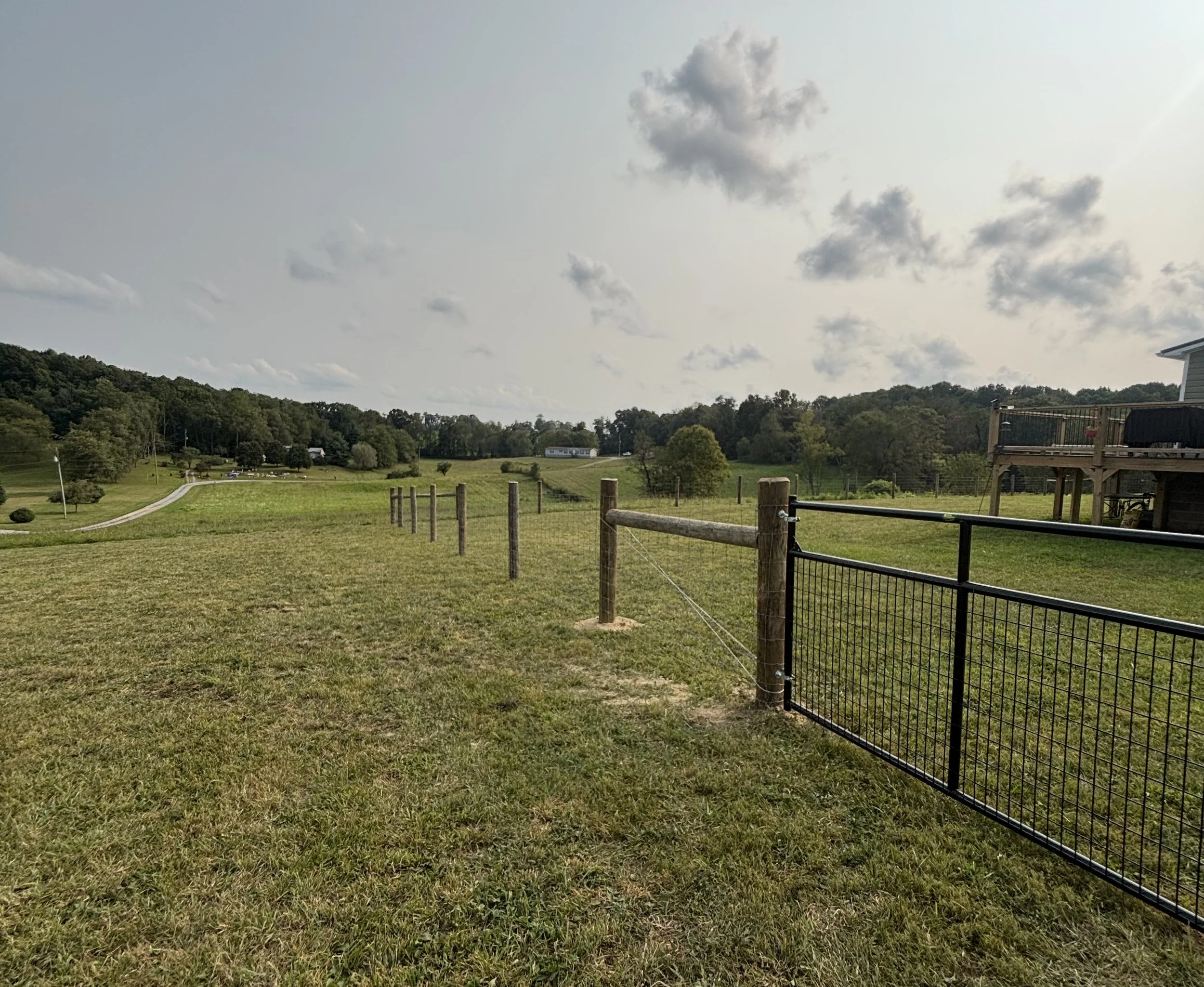 Open grassy field with a black metal gate and wooden fence in the foreground, rolling hills and trees in the background, partly cloudy sky, and a house with a deck on the right side.