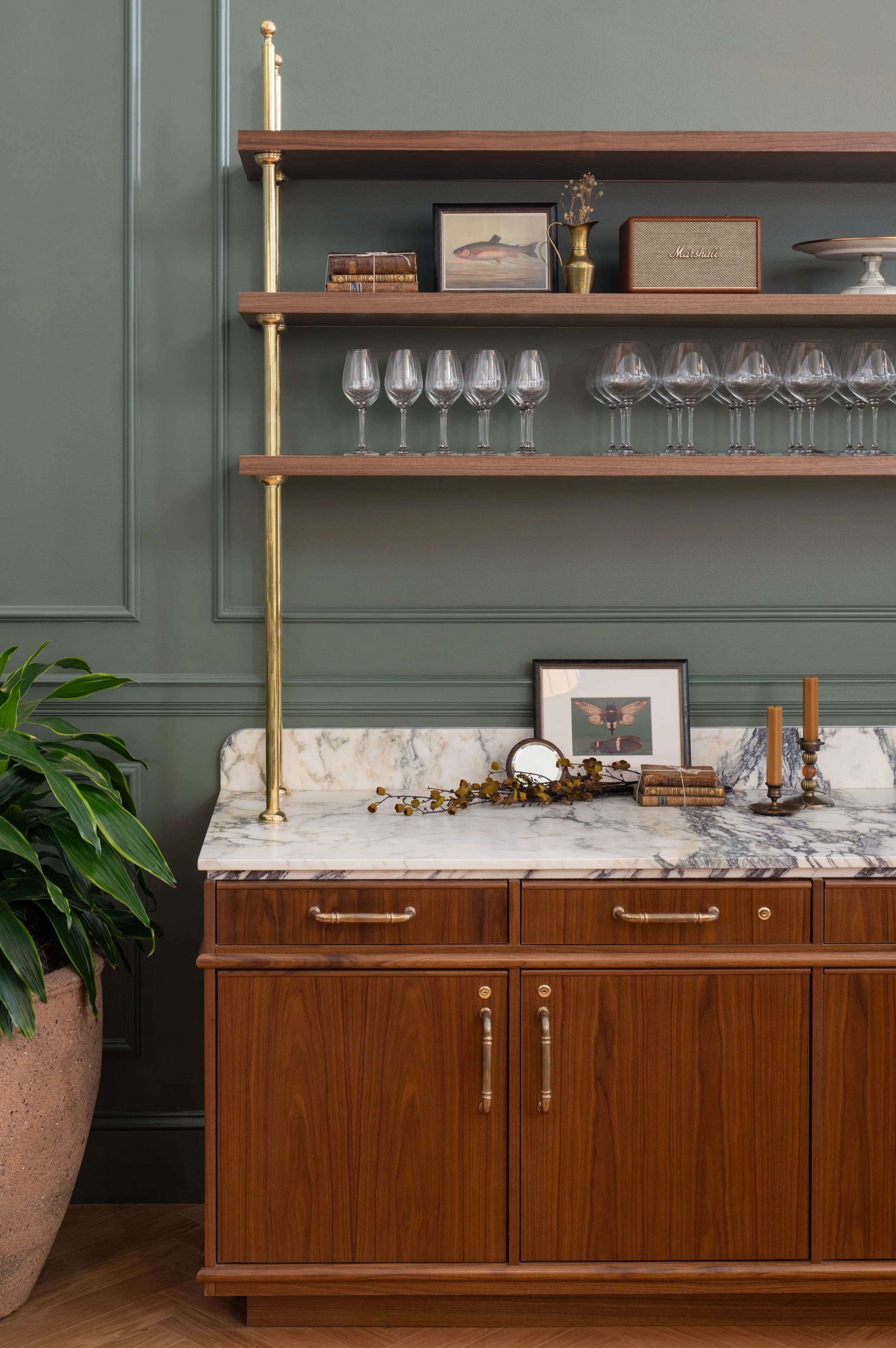 Elegant midcentury modern wooden sideboard with marble top, brass shelving, wine glasses, books, artwork, vase, Marshall speaker, candles, and houseplant.