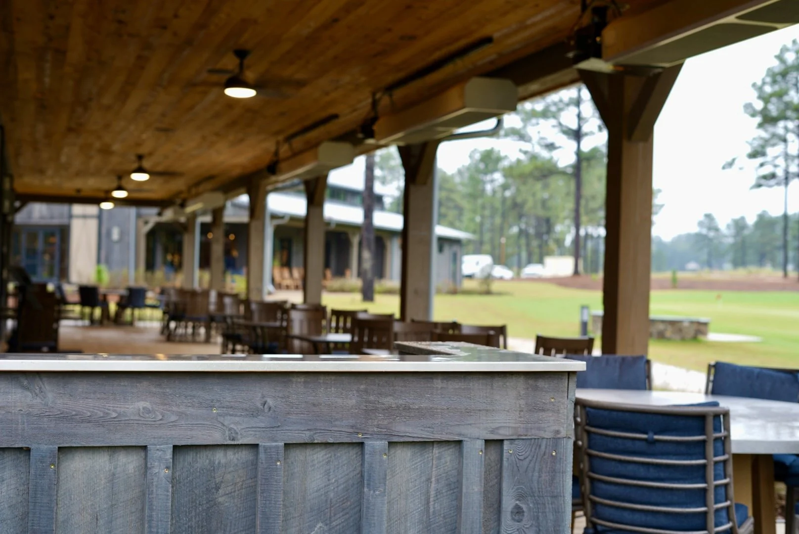 Covered outdoor restaurant patio with wooden bar, dining tables, blue chairs, and a pine-lined lawn