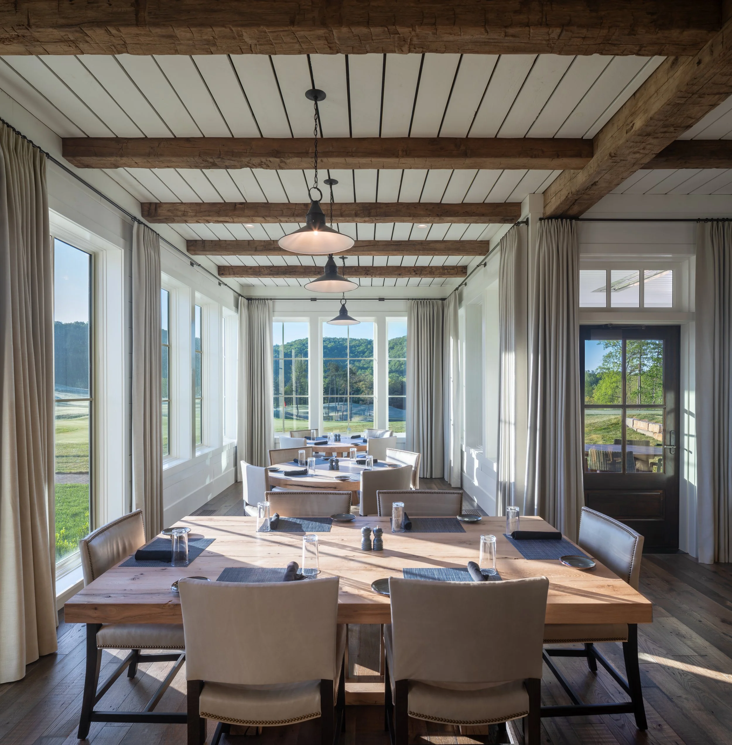 Bright rustic restaurant dining room with wooden tables, upholstered chairs, pendant lights, exposed beams, and scenic windows