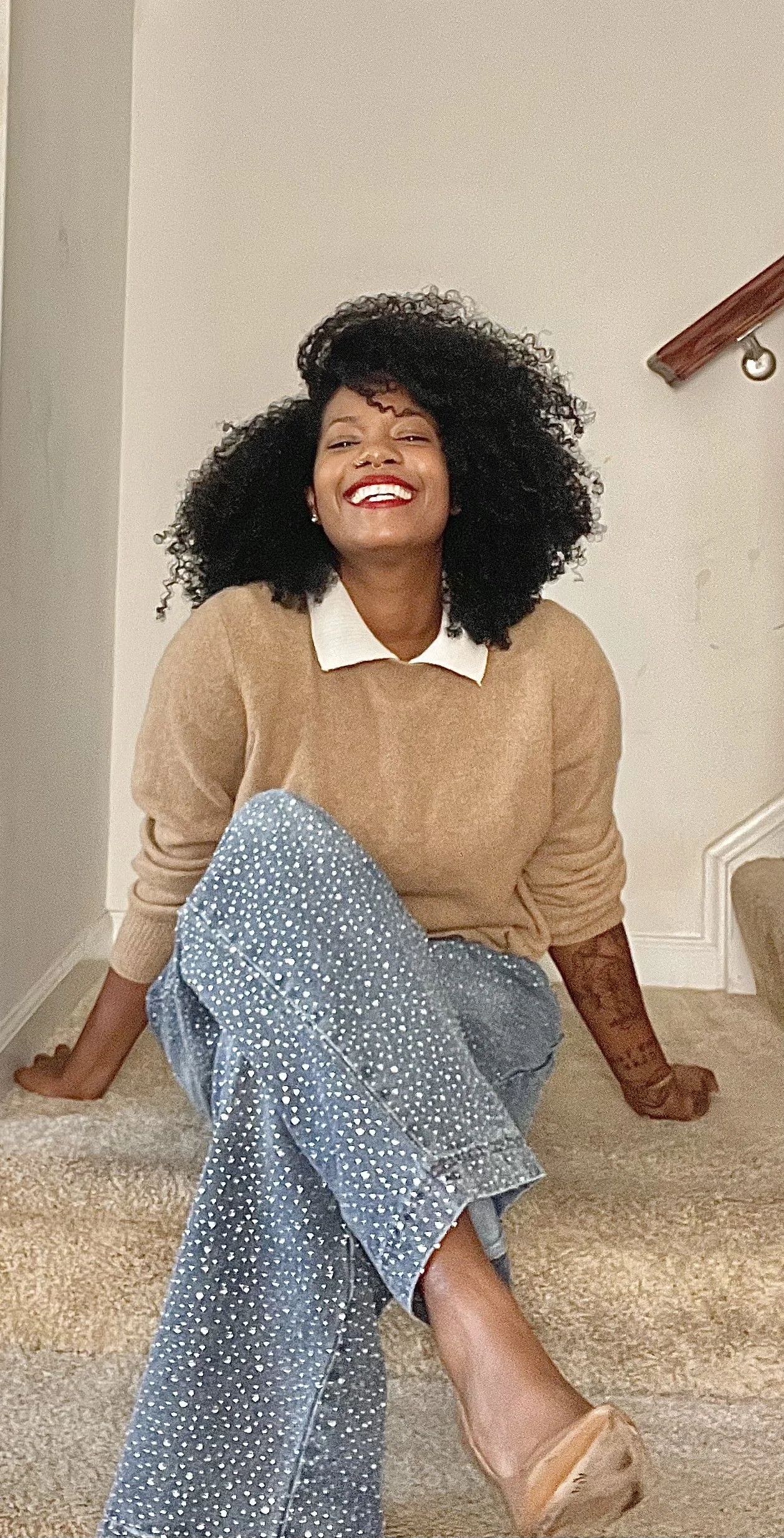 A woman with curly black hair sitting on a staircase, smiling, wearing a tan sweater with a white collar, polka dot pants, and tan shoes.