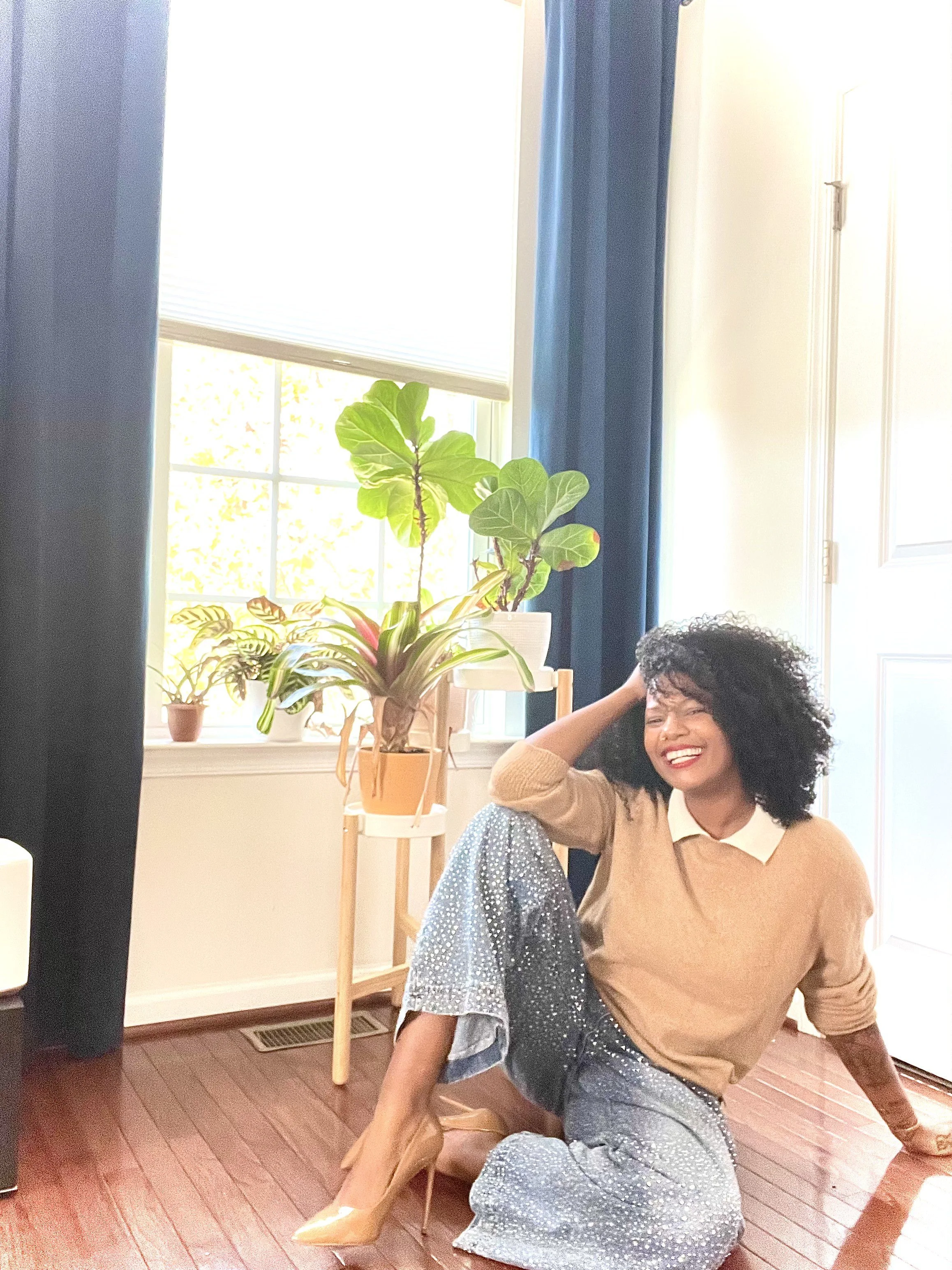 A woman with curly black hair sitting on a hardwood floor in a sunlit room, wearing a beige sweater, sparkly gray pants, and nude high heels, smiling and holding her head with one hand surrounded by green indoor plants near a window.