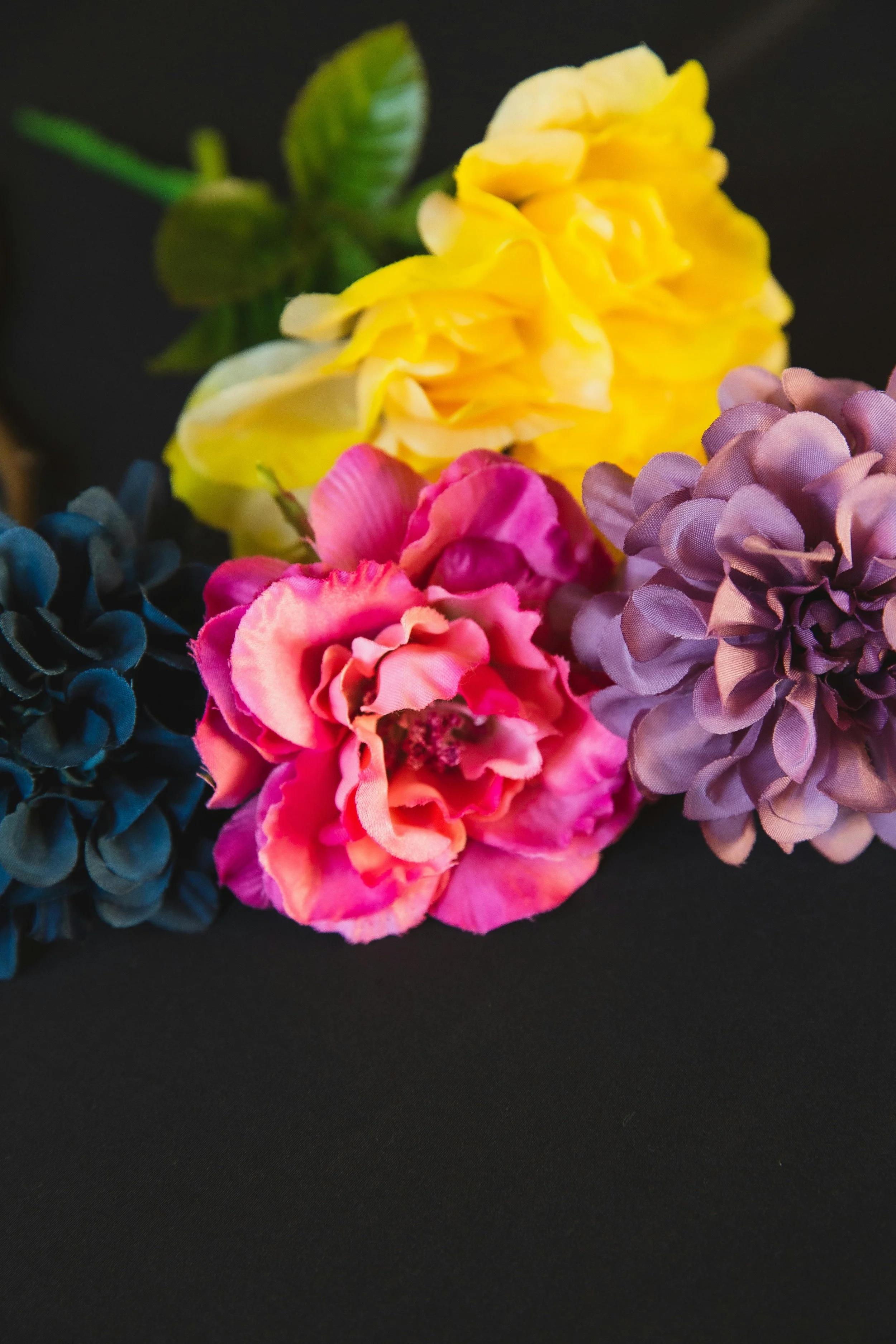 A bouquet of four artificial flowers: yellow, pink, purple, and dark blue, set against a black background.