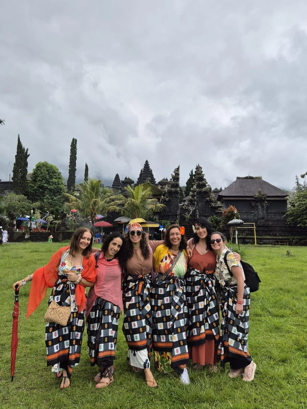 Grupo de seis mujeres en ropa tradicional, posando en un campo verde con un templo antiguo de fondo, bajo un cielo nublado en Bali.
