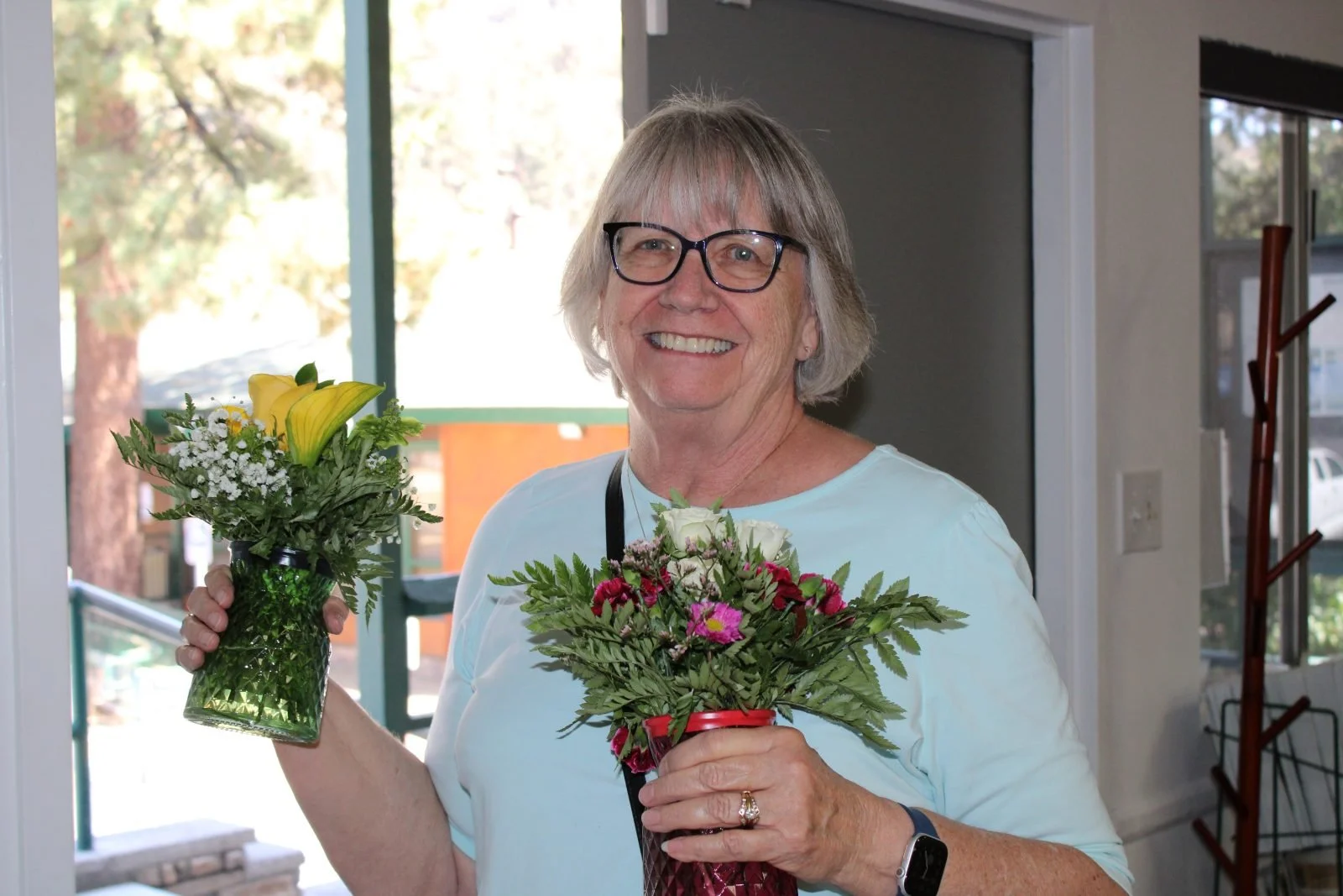 Smiling woman with glasses holding two flower bouquets, one with yellow lilies, the other pink and white flowers. Background of trees and a window.