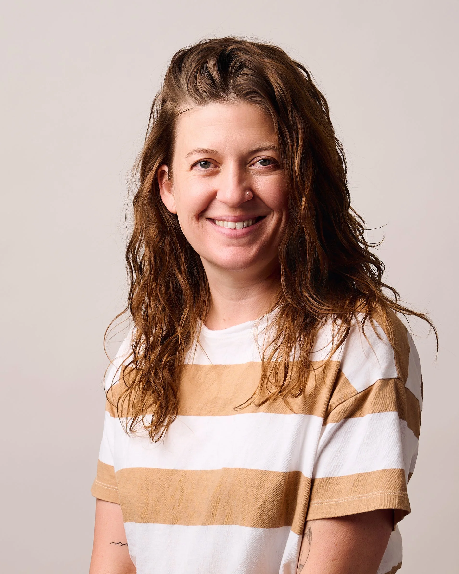 Erin DeGroot, a smiling woman with wavy brown hair wearing a white and tan striped T-shirt. Plain background. Warm, friendly expression. Casual, relaxed tone.