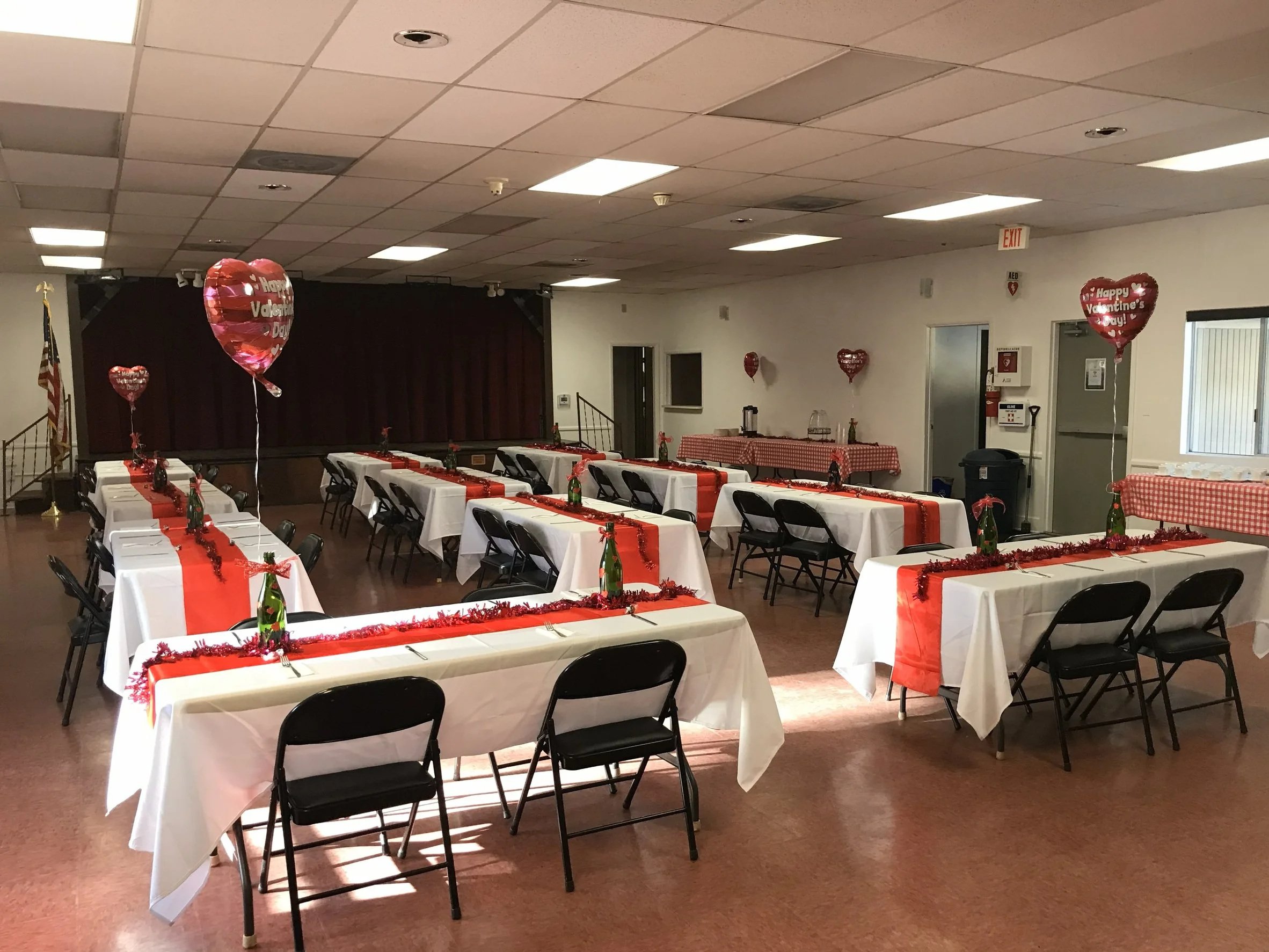 Spacious community building with shiny red-brown flooring, a small elevated stage with closed red curtains, and two side tables. 