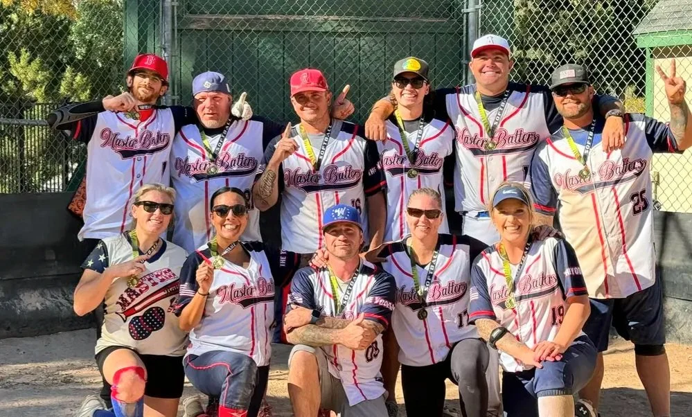 A joyful softball team poses for a group photo in uniforms, holding medals. Smiling faces convey pride and camaraderie. Chain-link fence and trees in the background.