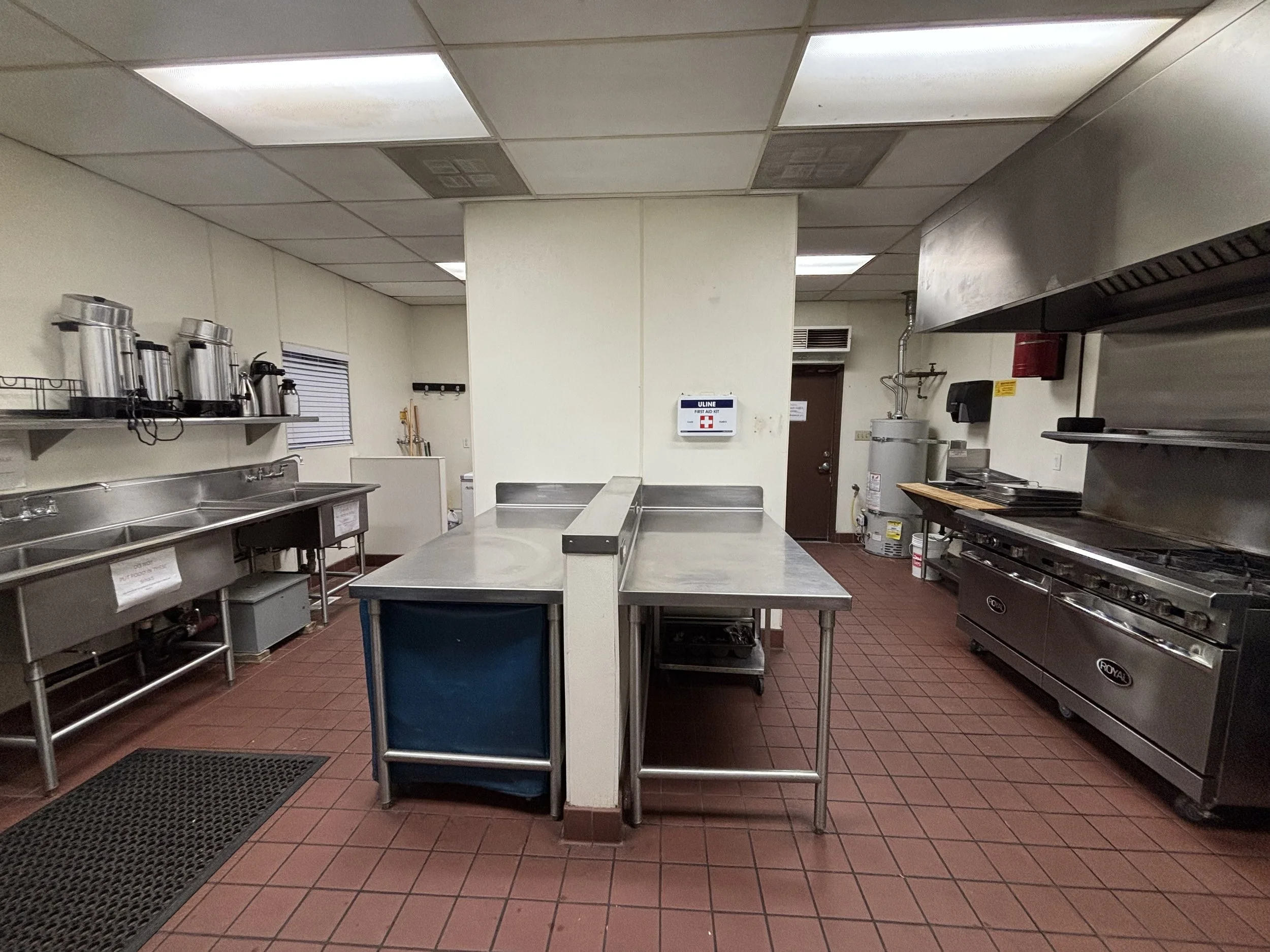 Community commercial kitchen with stainless steel appliances, including a large stove and oven. Brick floor, metal counters, and visible water heater.