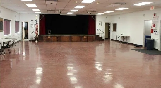 Spacious community building with shiny red-brown flooring, a small elevated stage with closed red curtains, and two side tables. 