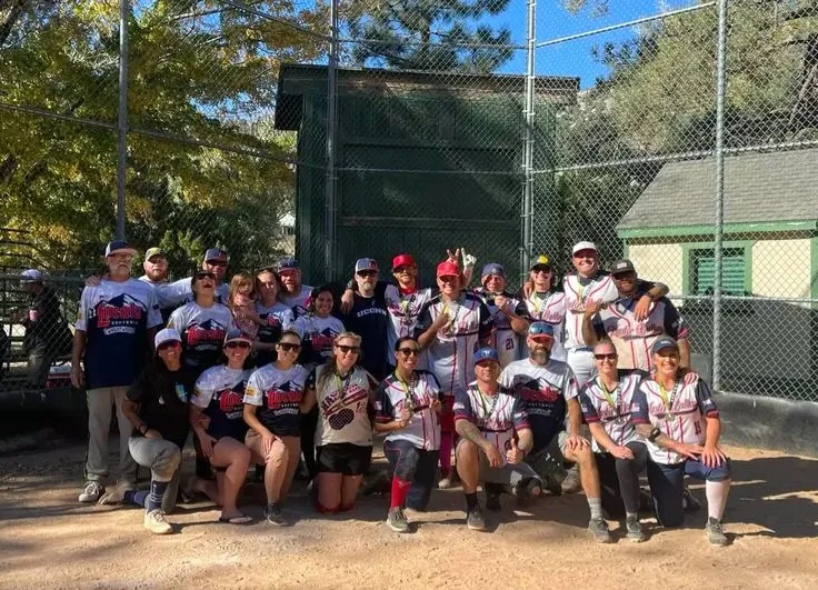 A group of softball players in matching uniforms pose on a field. They're smiling and celebratory, with a sunny sky and trees in the background.