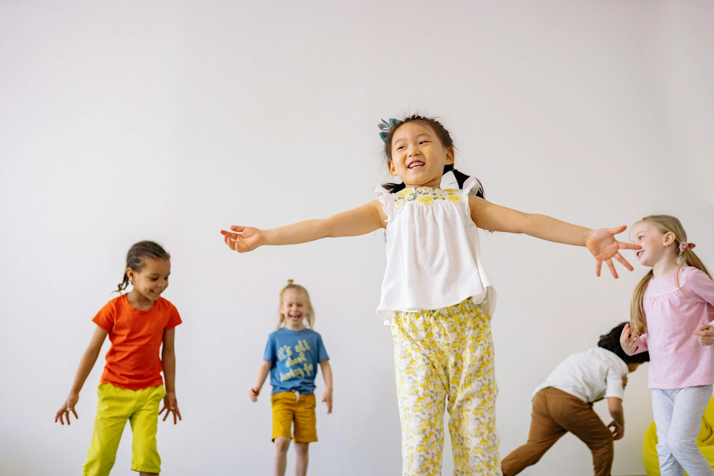 Children joyfully dance in a bright room. A girl in a white top and yellow pants leads, arms wide and smiling. Others in colorful outfits follow, laughing.