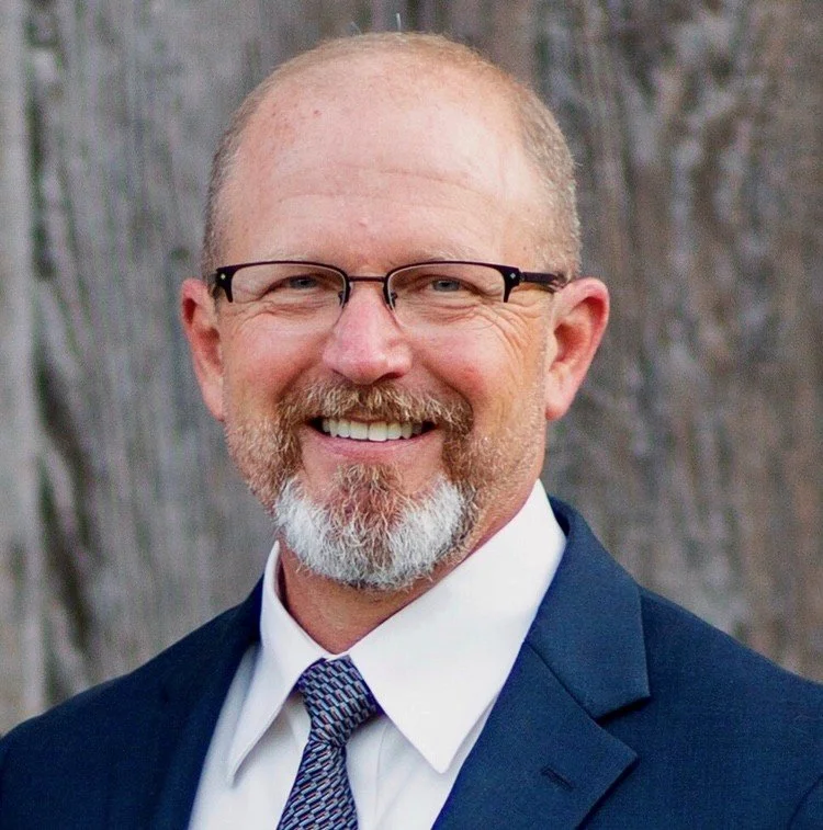Dave Schoenwetter, a smiling man with glasses, a beard, and a mustache, wears a dark blue suit, white shirt, and patterned tie, standing in front of a wood-textured background.