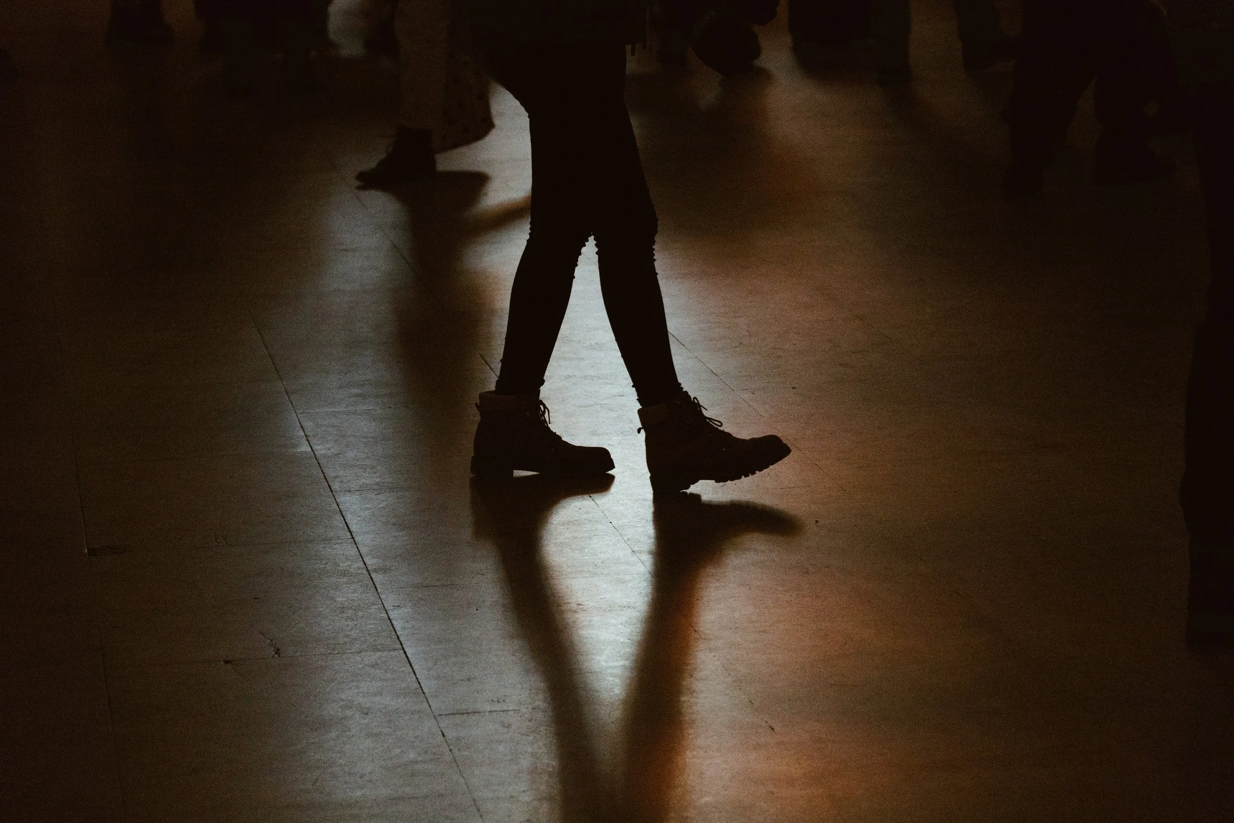 Silhouetted legs in boots walking on a shiny floor cast long shadows in a dimly lit space, conveying a sense of movement and mystery.