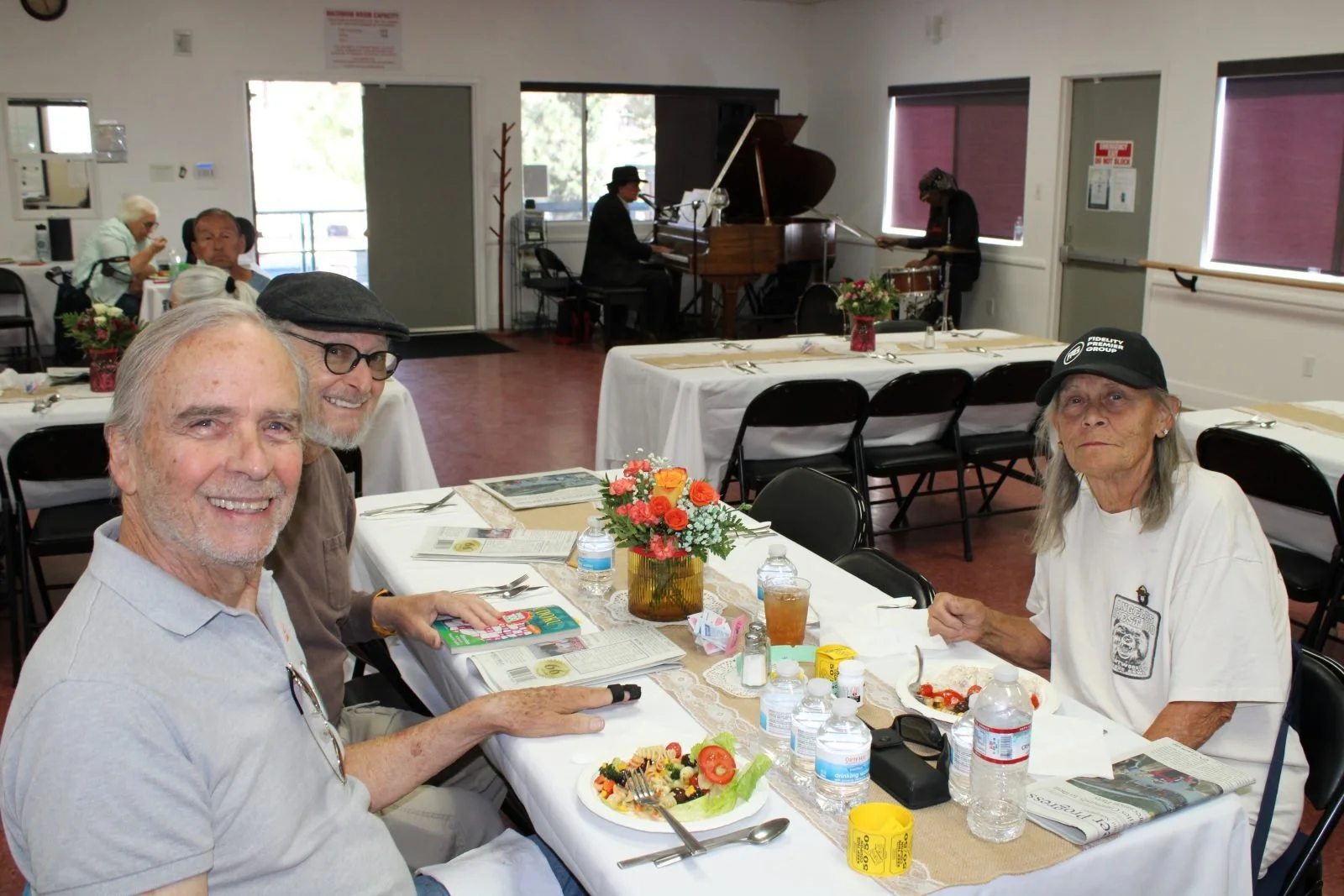 Three seniors sit at a dining table with colorful salads and drinks, smiling. A pianist plays in the background. The atmosphere is warm and cheerful.