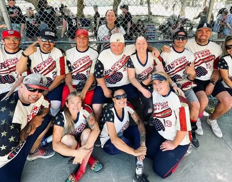 A joyful softball team poses for a group photo on a sunny day. Ten players wear matching patriotic jerseys, sitting and kneeling in front of a chain-link fence.